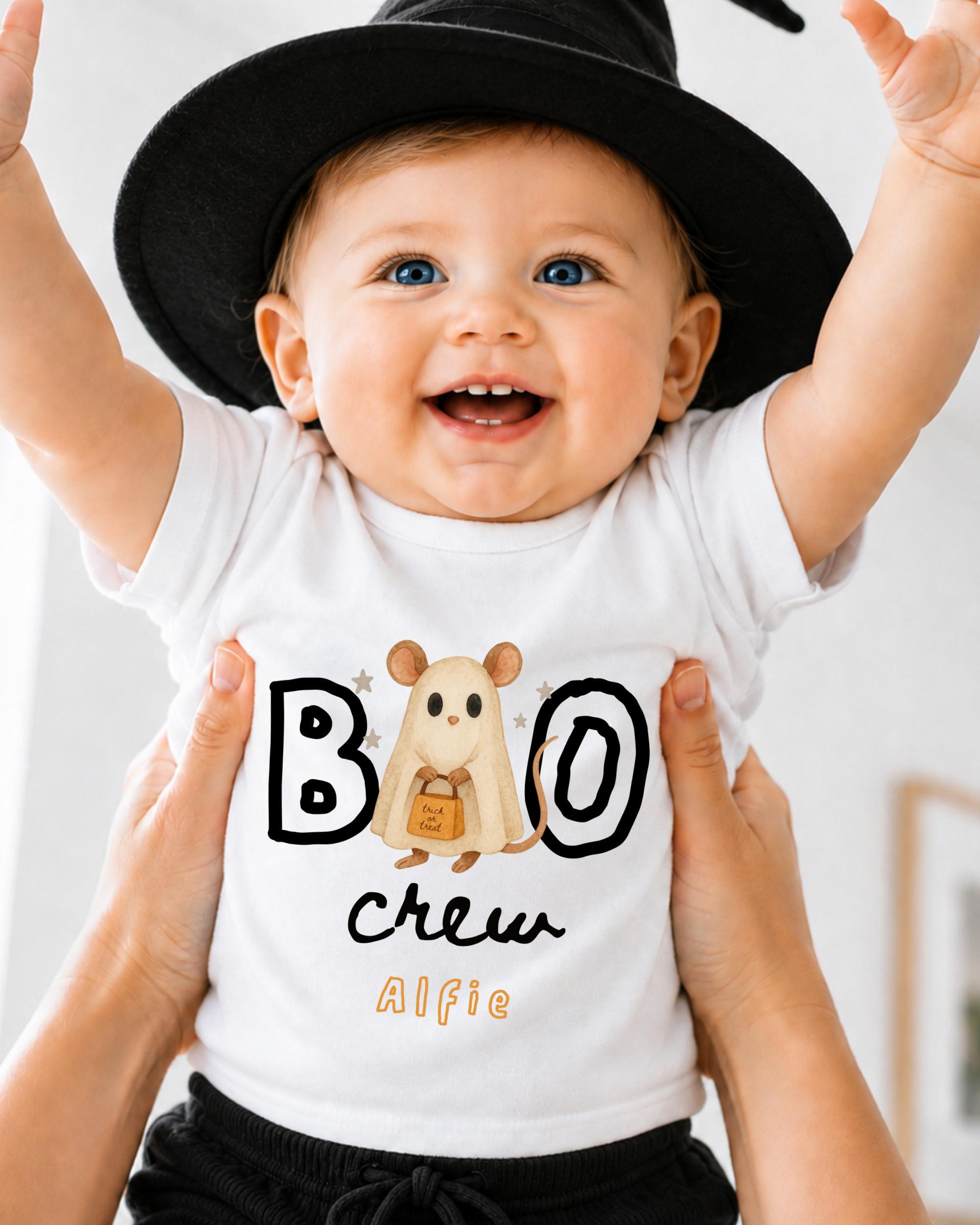 A smiling baby wearing a white t-shirt with a cartoon character and the text "BOO crew Alfie" is being held up by someone's hands.