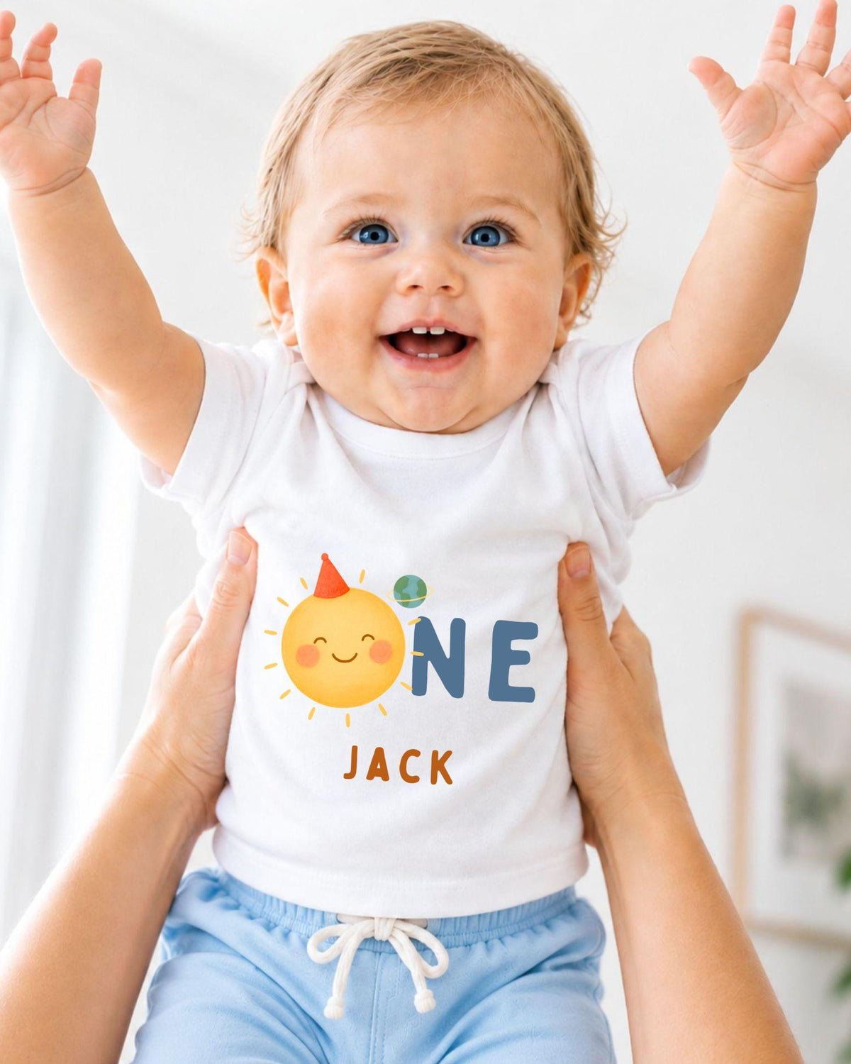 A smiling baby wearing a white shirt with a sun and name printed on it, being held up by someone's arms.