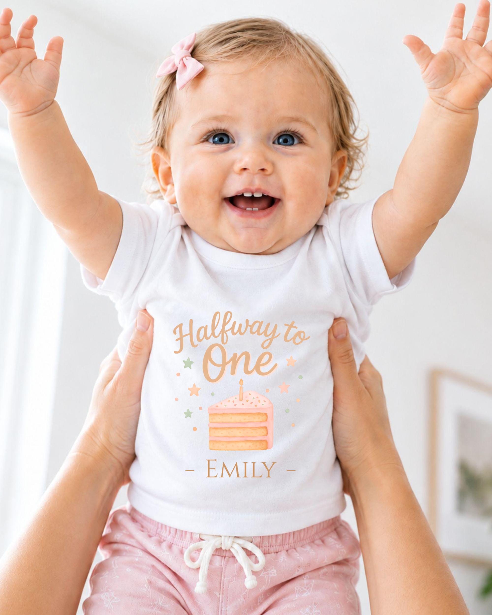 A smiling baby girl wearing a white t-shirt with a birthday cake design and the text "Halfway to One - Emily" is being held up by someone's arms.