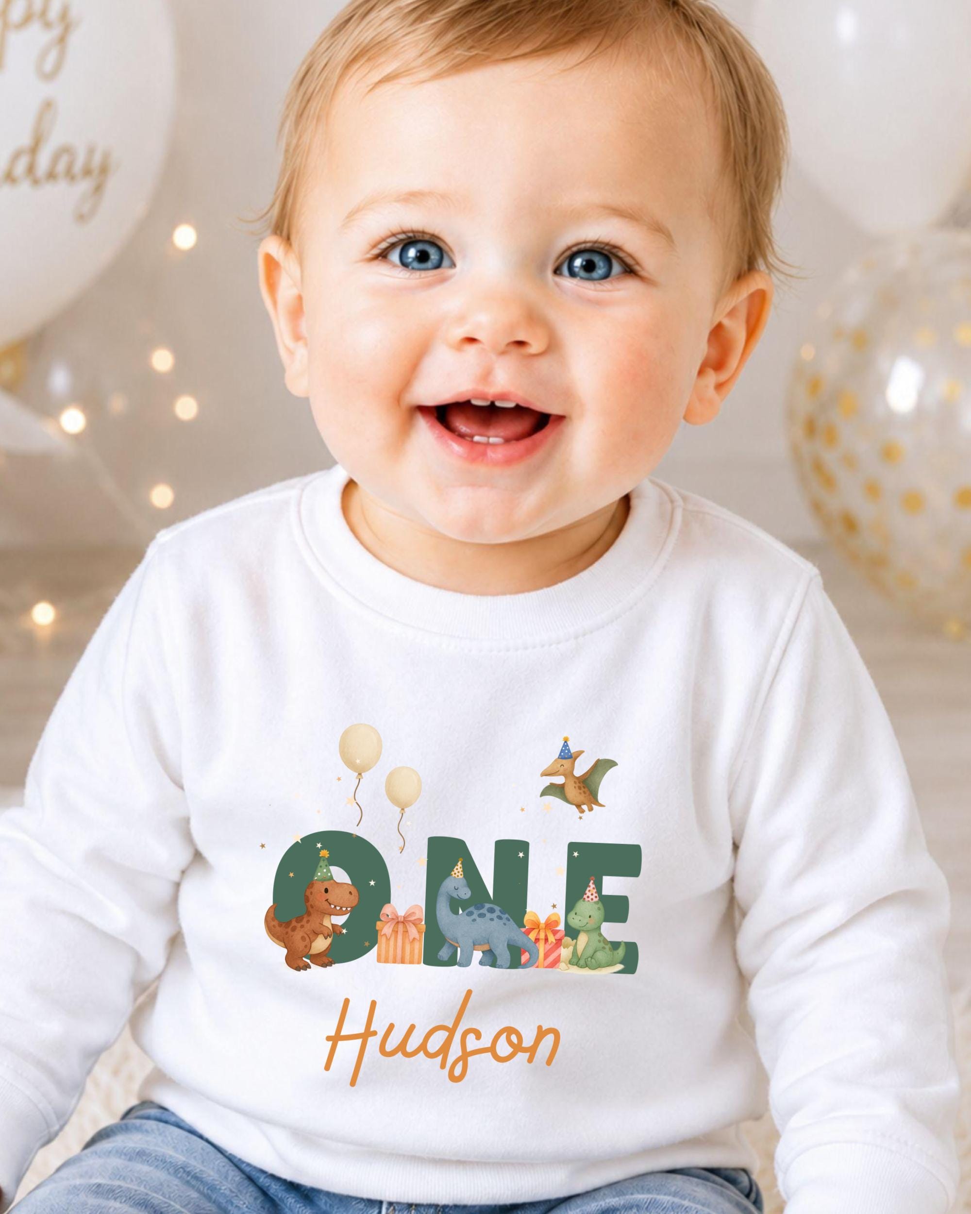 A smiling baby boy wearing a white shirt with the word "One" and a dinosaur graphic, sitting in front of birthday balloons.