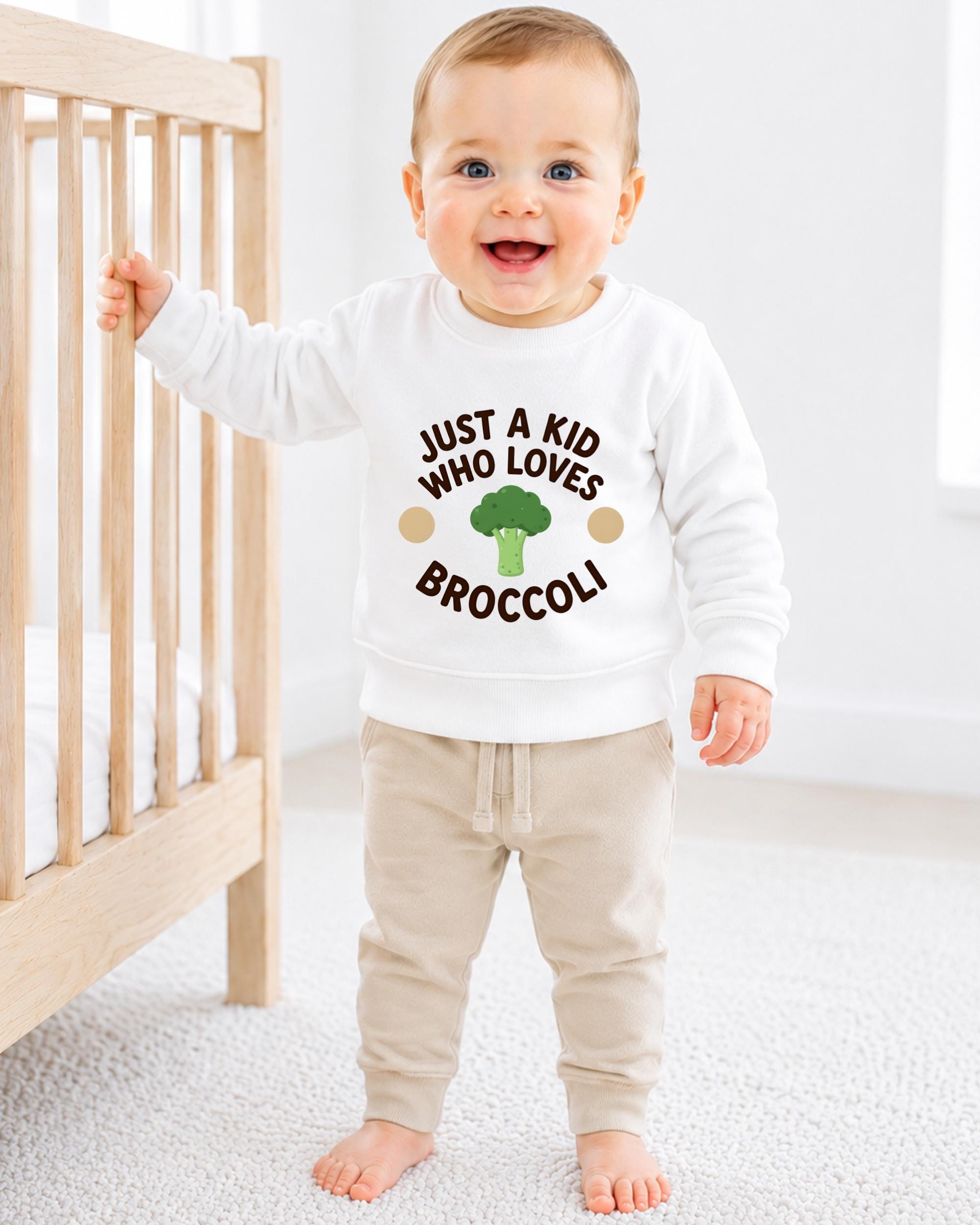 A smiling baby boy wearing a white sweatshirt with the text "JUST A KID WHO LOVES BROCCOLI" stands in front of a wooden crib.