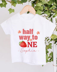 A white t-shirt with a printed design on it, hanging on a wooden hanger. The design features a strawberry and the text "half way to one" along with the name "Sophia".