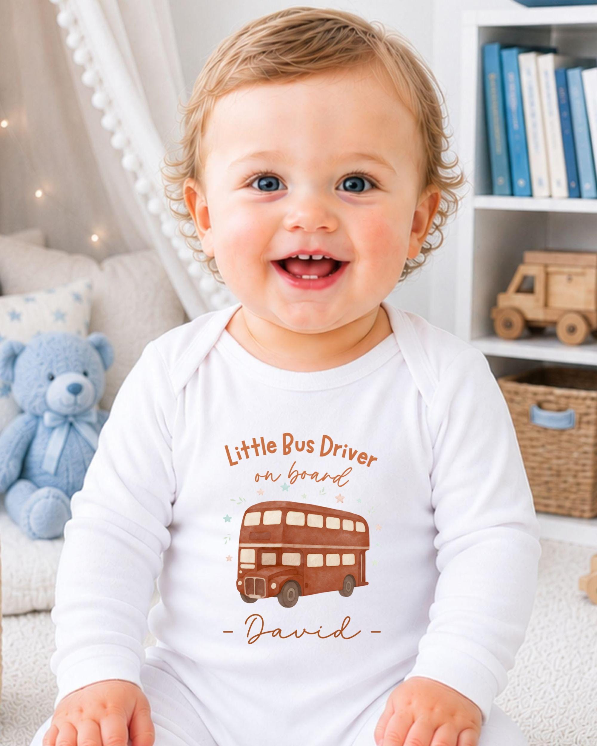 A smiling baby boy wearing a white onesie with a double-decker bus design and the text "Little Bus Driver on board David" printed on it.