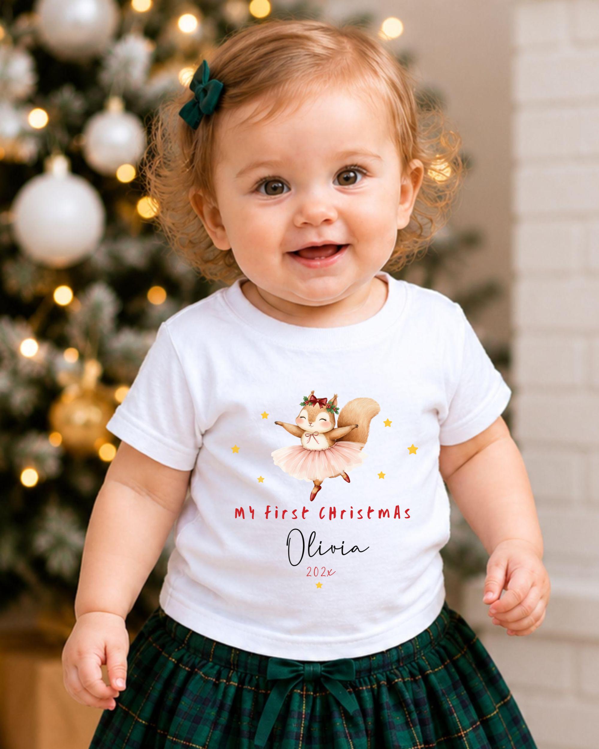 A smiling baby girl wearing a white t-shirt with a cartoon squirrel and the text "My first Christmas Olivia" printed on it, standing in front of a Christmas tree.