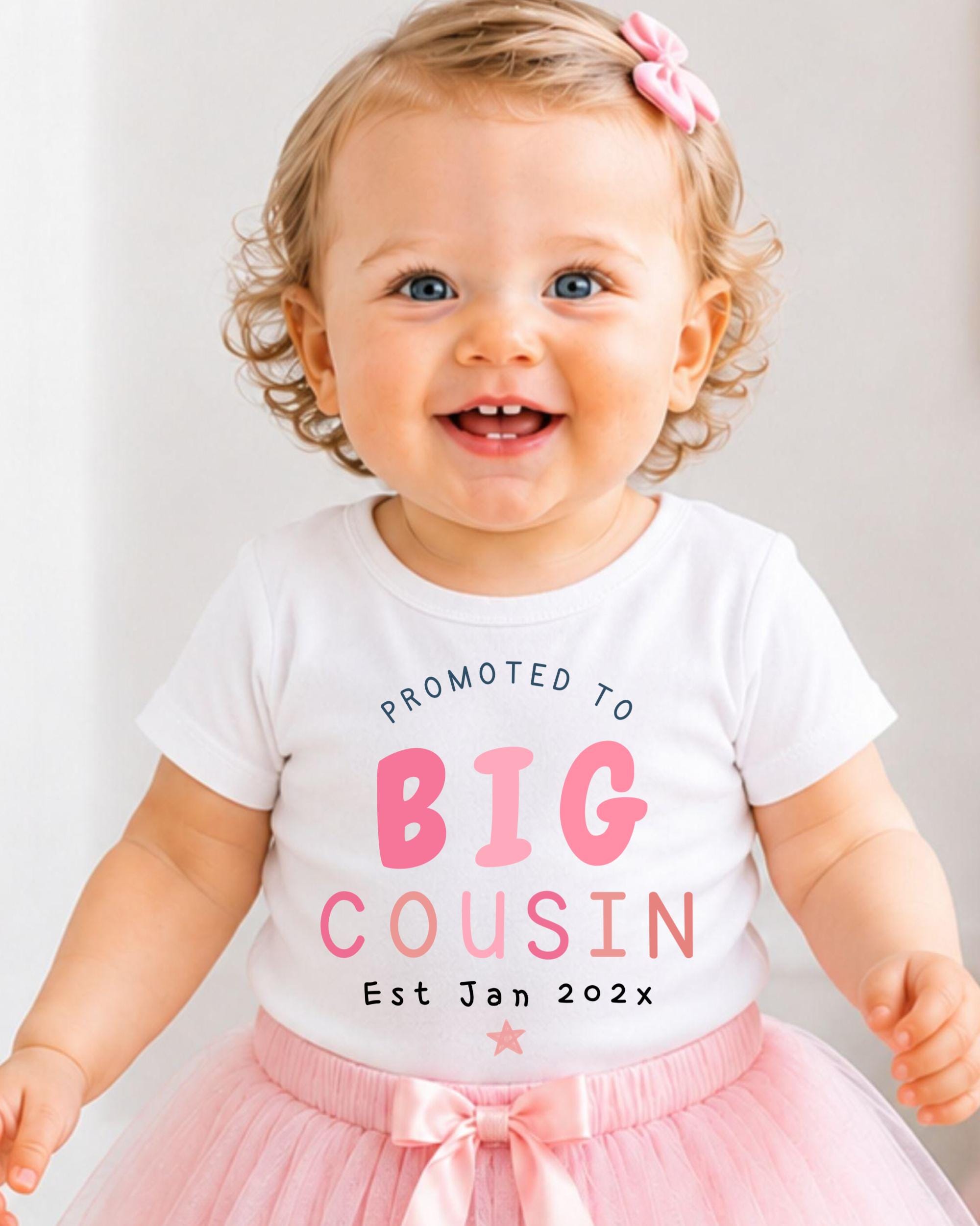 A smiling baby girl wearing a white t-shirt with text promoting a big cousin and a pink tutu.