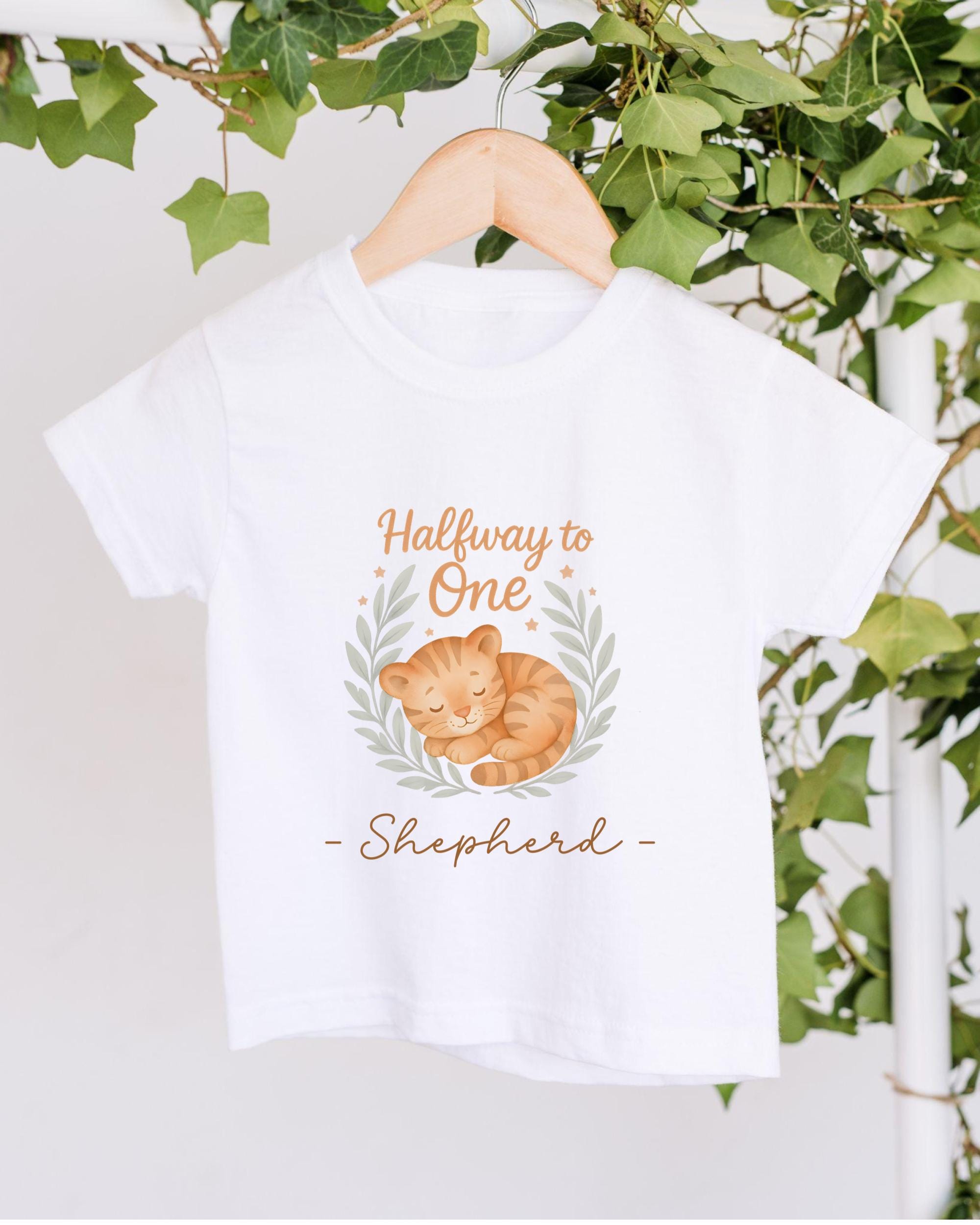A white t-shirt with a graphic design of a sleeping cat and the text "Halfway to One Shepherd" printed on it, hanging on a wooden hanger.