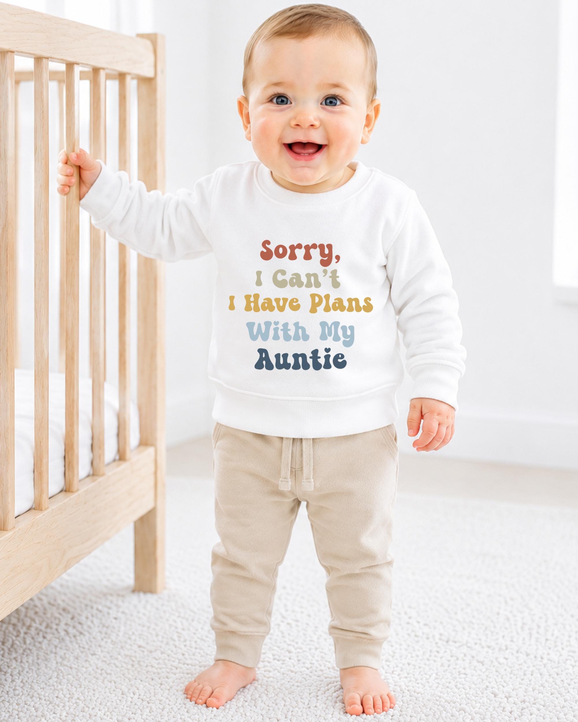 A smiling baby boy wearing a white sweatshirt with text printed on it, standing in front of a wooden crib.