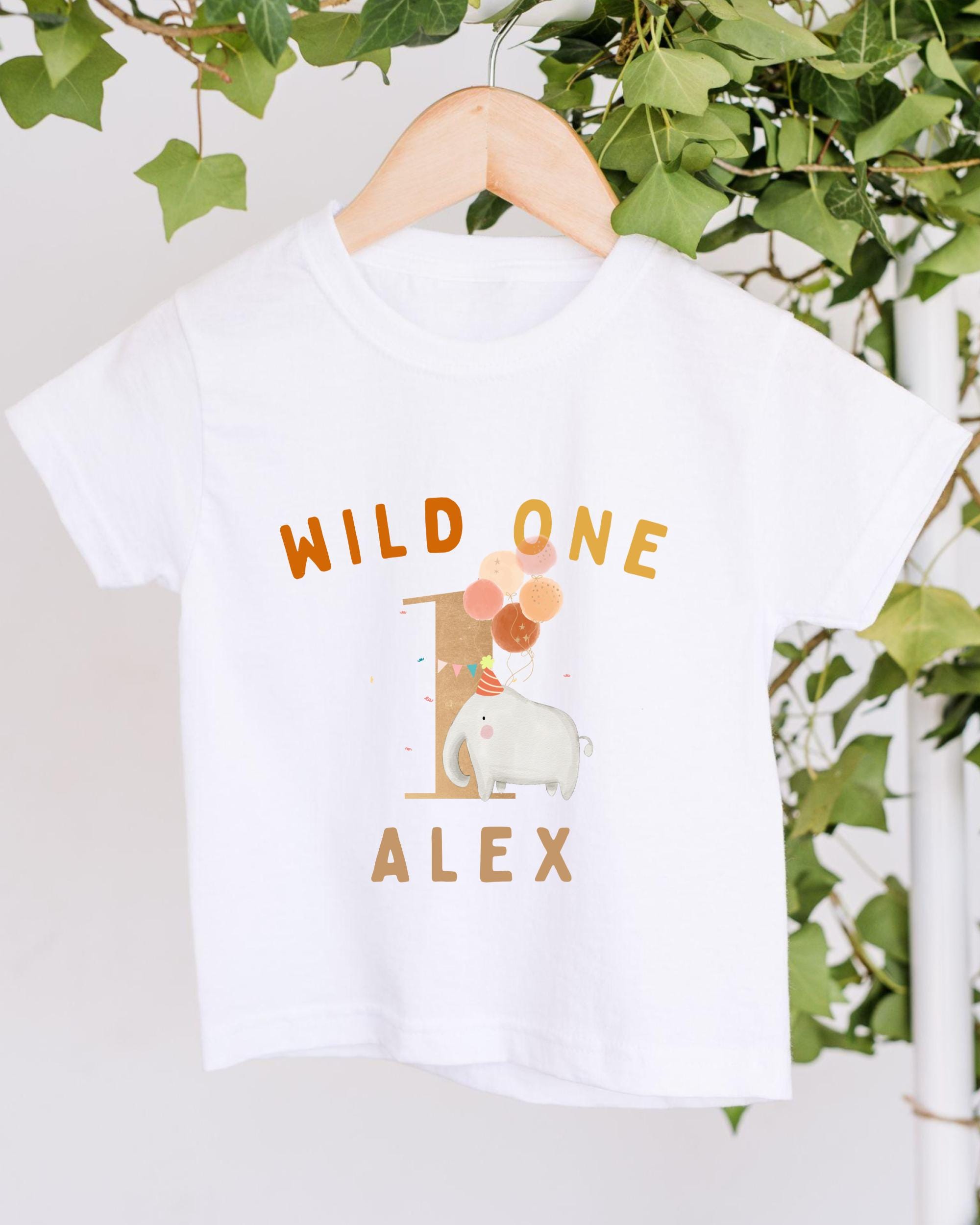 A white t-shirt with a printed design on it, hanging on a wooden hanger. The design features a white elephant surrounded by colorful balloons, with the text "WILD ONE ALEX" printed on the shirt.
