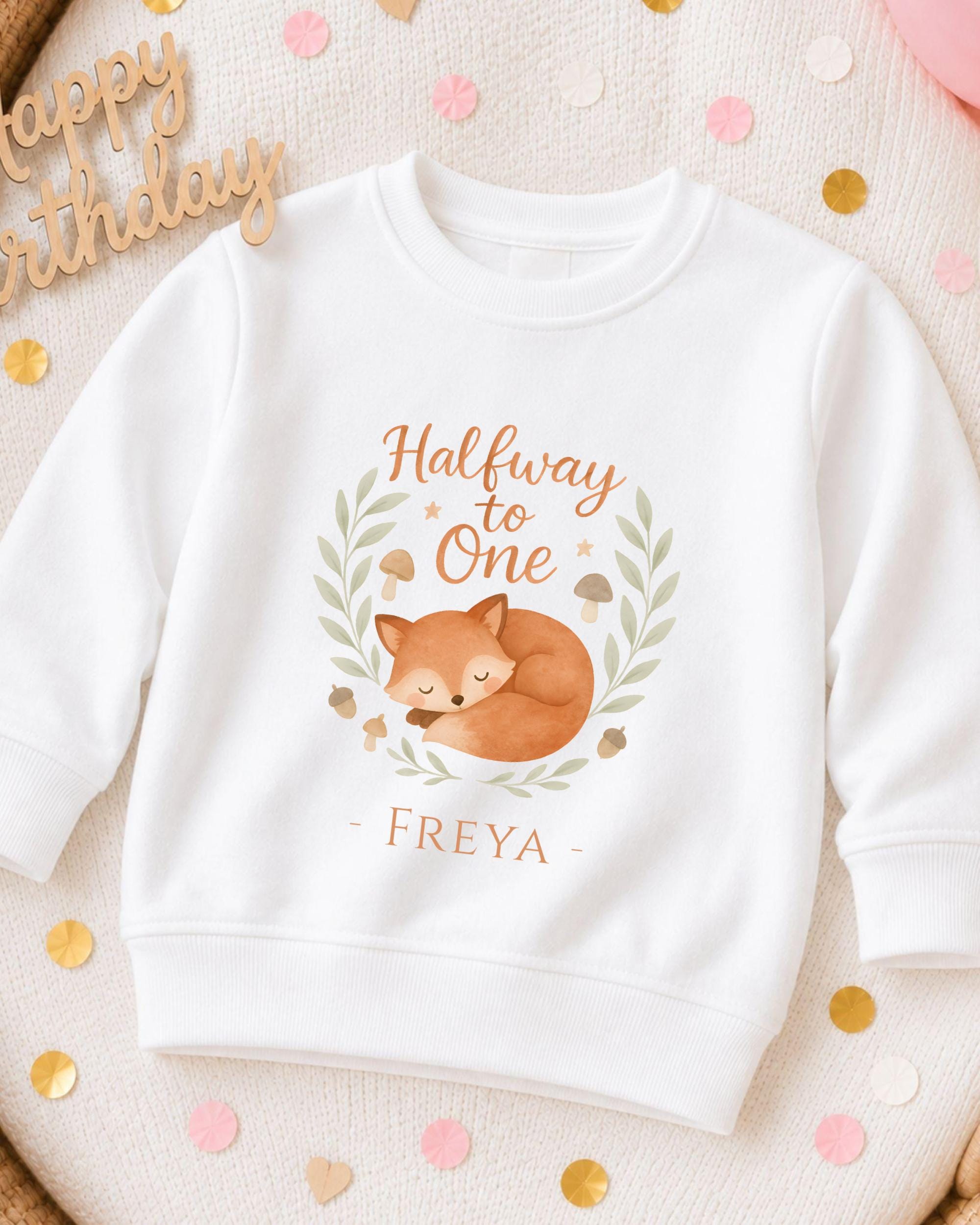 A white sweatshirt with a graphic design featuring a sleeping fox and the text "Halfway to One" and "Freya" printed on it.
