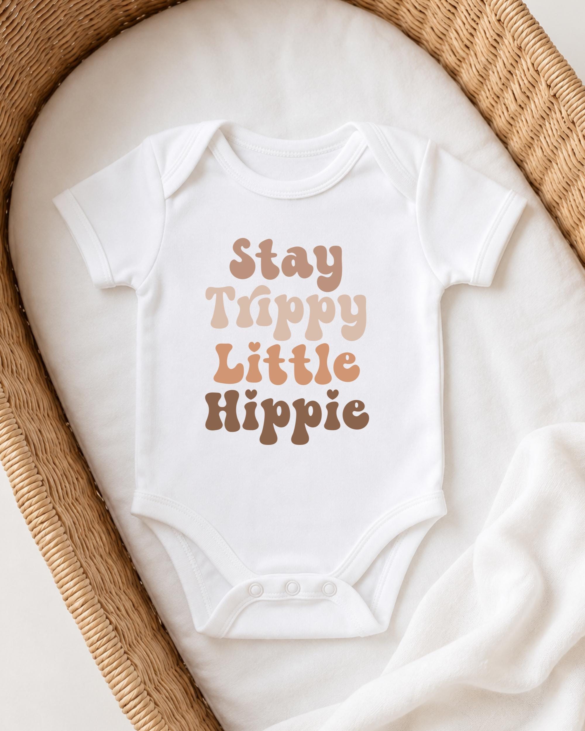 A white baby onesie with the text "Stay Trippy Little Hippie" printed on it, laid out on a white blanket in a wicker basket.