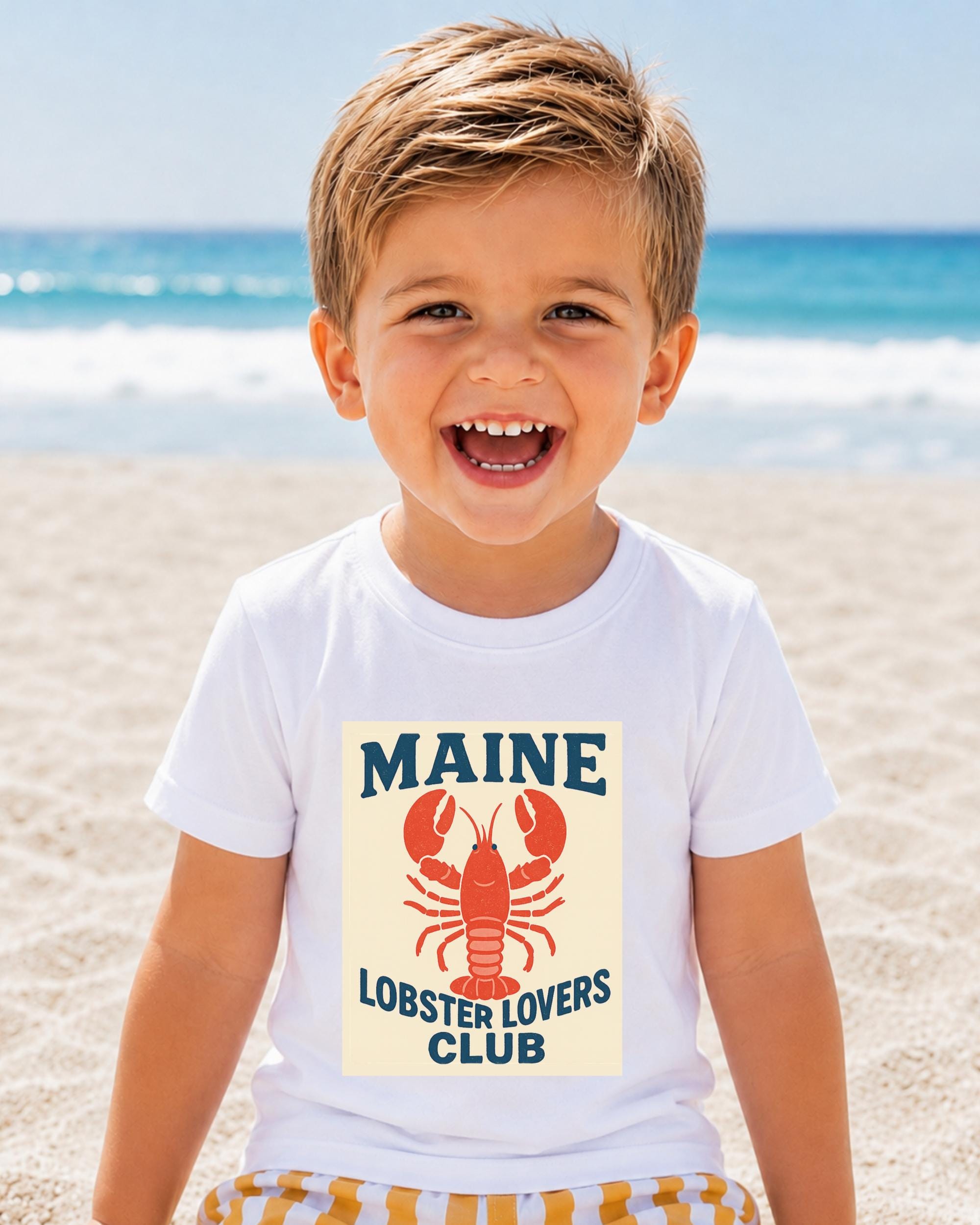 A young boy with a big smile on his face, wearing a white t-shirt with a lobster graphic and text, standing on a sandy beach with the ocean in the background.