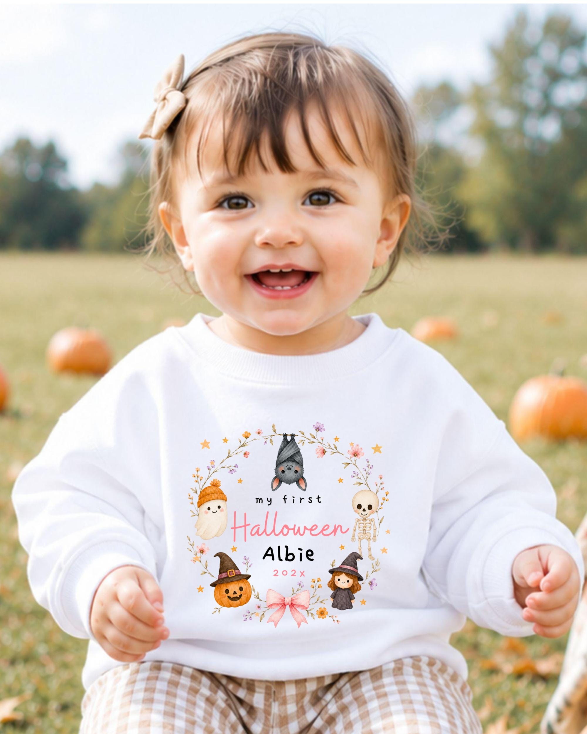A smiling baby girl wearing a white sweater with a Halloween-themed design and text.