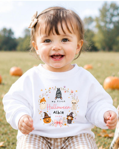 A smiling baby girl wearing a white sweater with a Halloween-themed design and text.