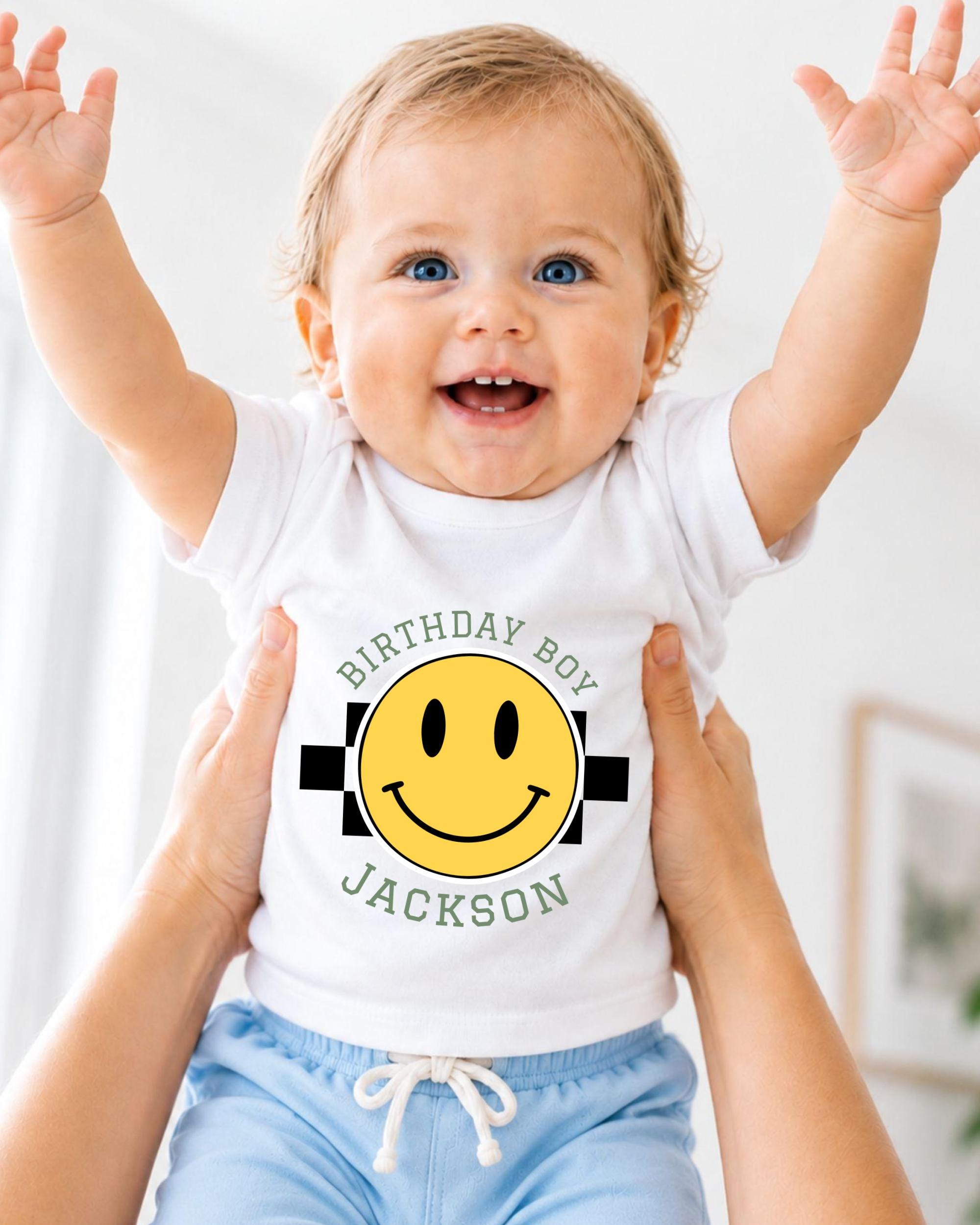 A smiling baby wearing a white t-shirt with a yellow smiley face and the text "Birthday Boy Jackson" is being held up by someone's arms.