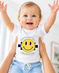 A smiling baby wearing a white t-shirt with a yellow smiley face and the text "Birthday Boy Jackson" is being held up by someone's arms.