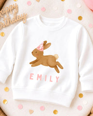 A white sweatshirt with a cartoon rabbit wearing a party hat and the name "EMILY" printed on it, surrounded by confetti.