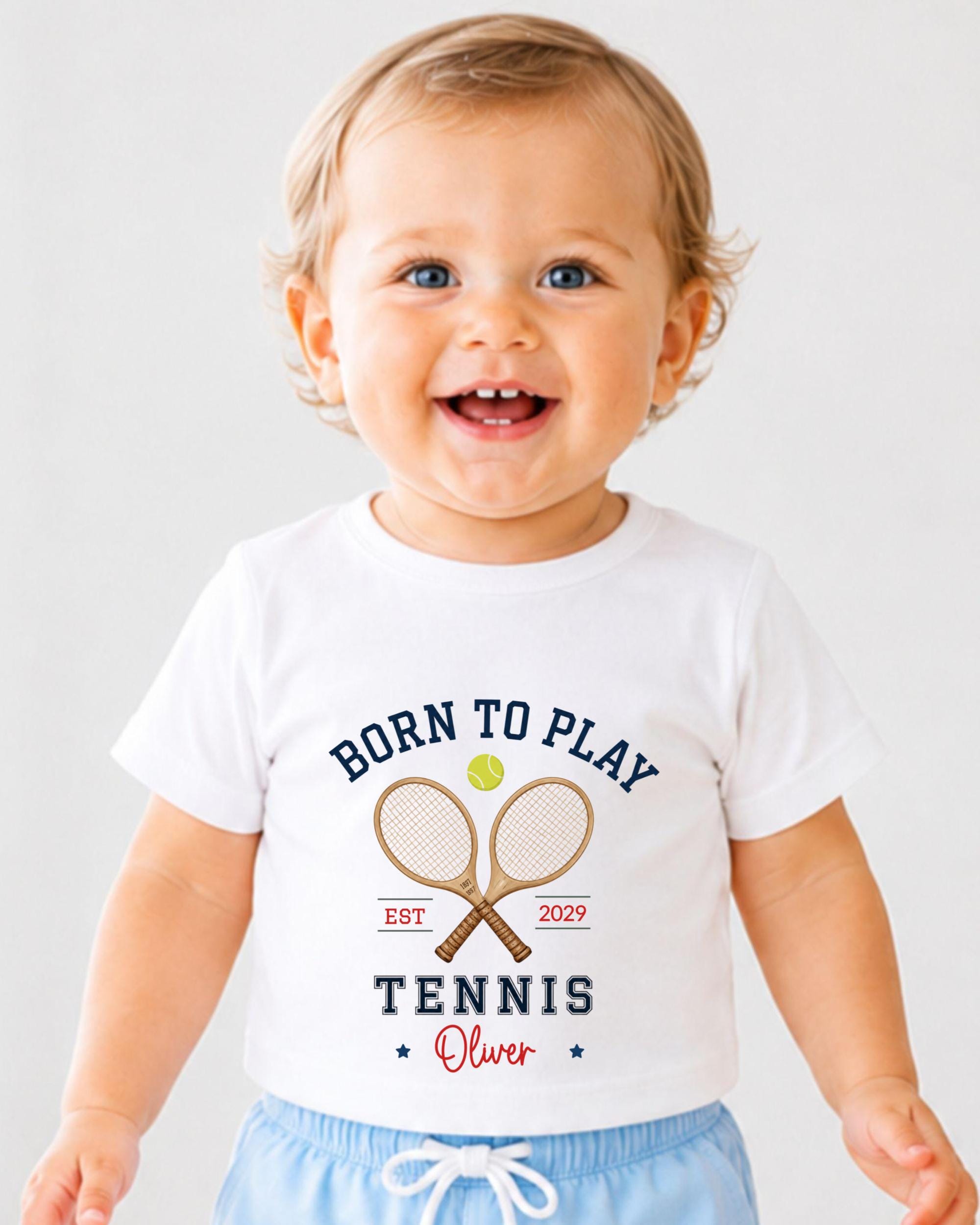 A smiling baby boy wearing a white t-shirt with a tennis racket and ball graphic, along with the text "Born to play tennis Oliver" printed on it.