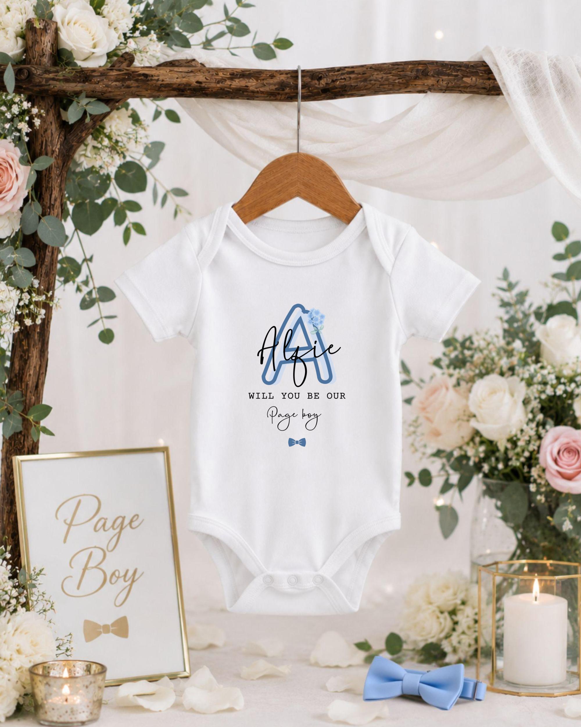 A baby onesie with the text "Will you be our Page Boy?" is hanging on a wooden hanger, surrounded by floral decorations and a candle.