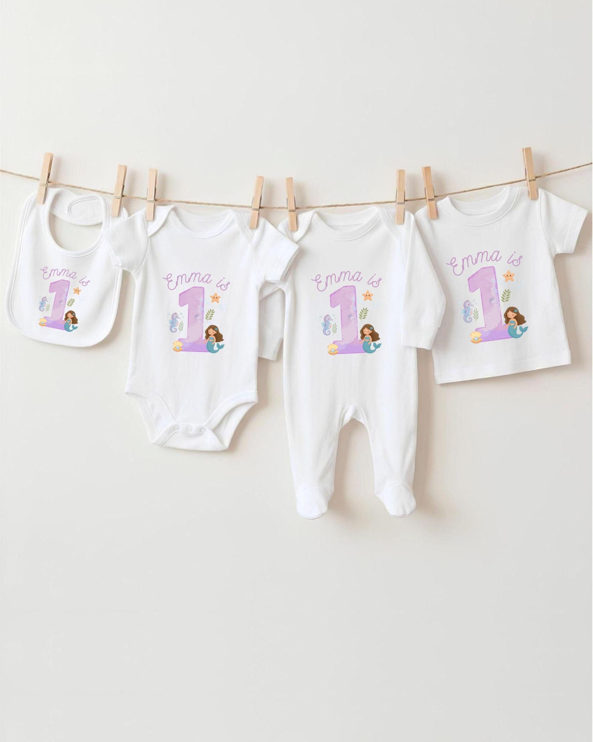 A collection of white baby clothes, including onesies and pajamas, with the name 'Emma' printed on them, along with a mermaid image. The clothes are hanging on a clothesline.