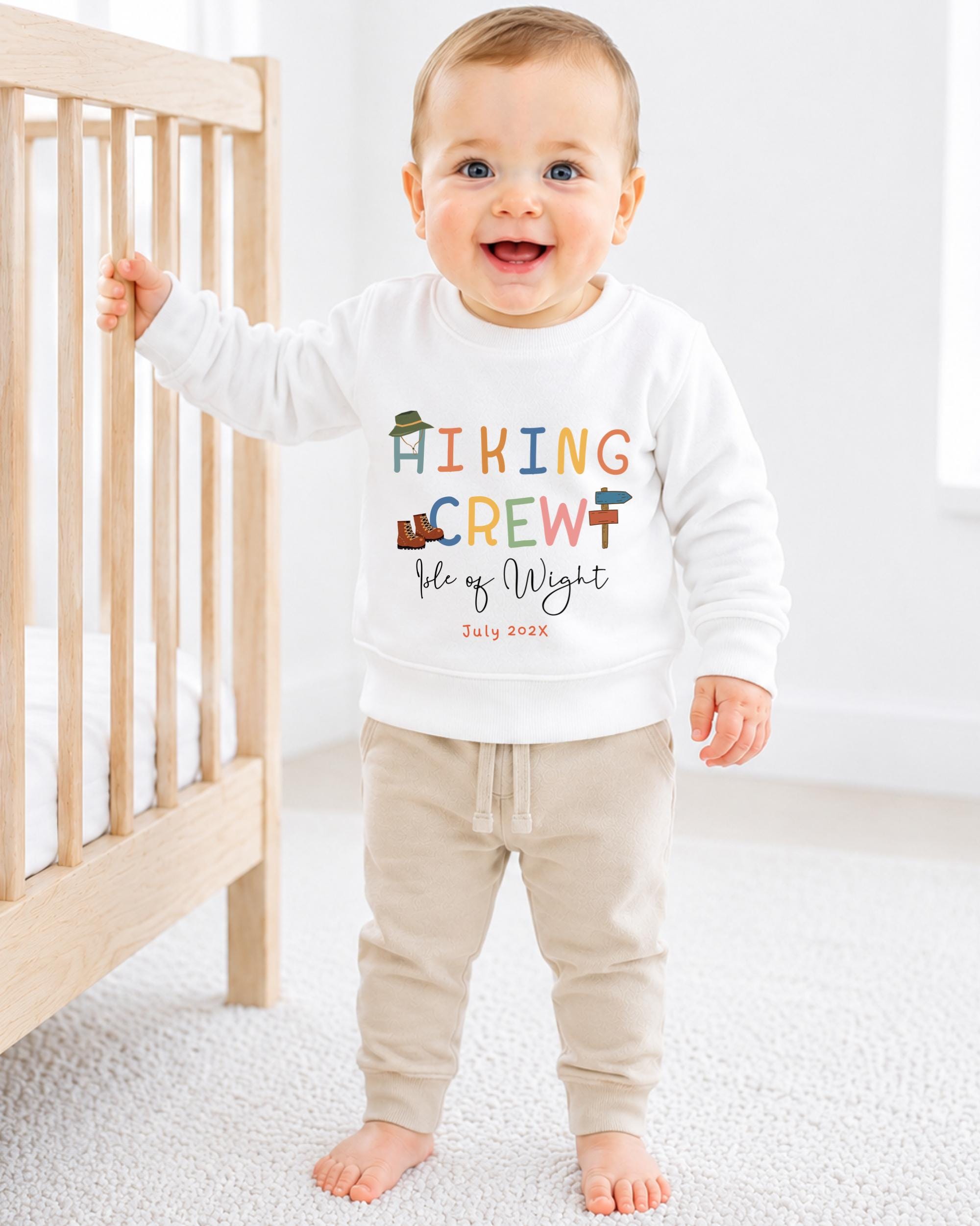 A smiling baby boy wearing a white sweatshirt with the text "HIKING CREW" and "Fleece of Wight" printed on it, standing in front of a wooden crib.