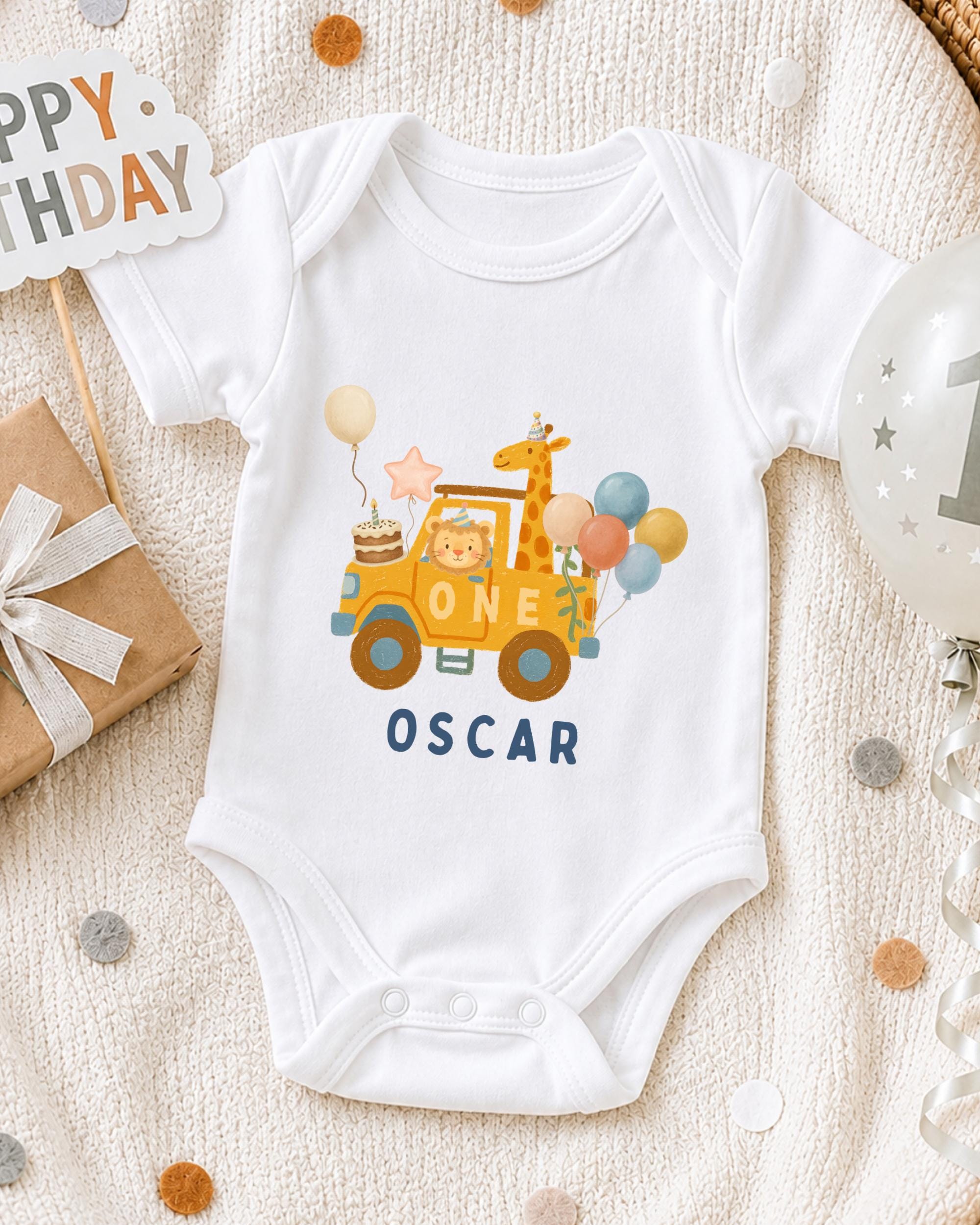 A white onesie with a cartoon car and a giraffe on it, along with a "Happy Birthday Oscar" sign, is displayed on a bed with confetti.