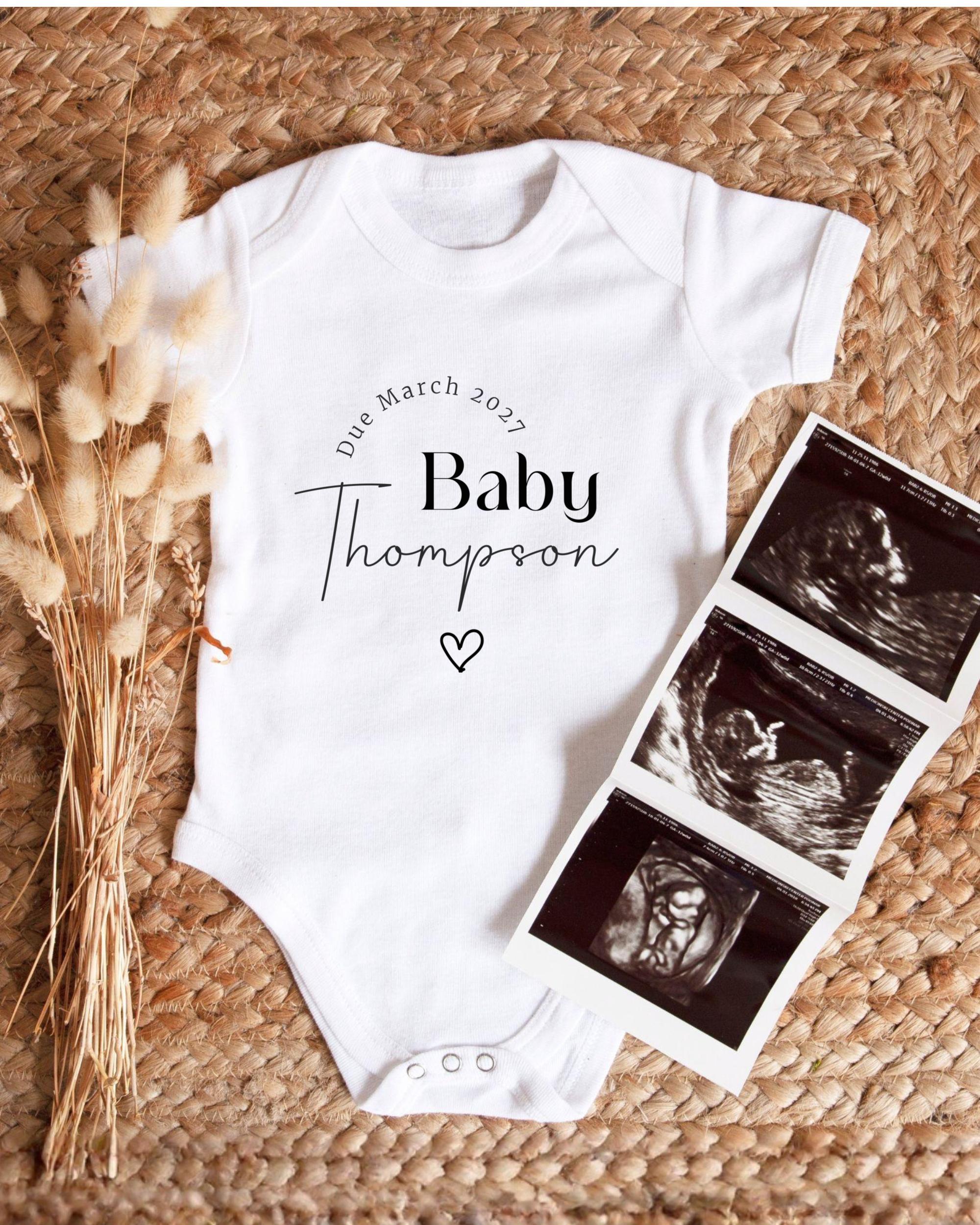 A white baby onesie with the text "Die March 2027 Baby Thompson" printed on it, along with a heart symbol. The onesie is placed next to a folded photograph of an ultrasound image of a baby.