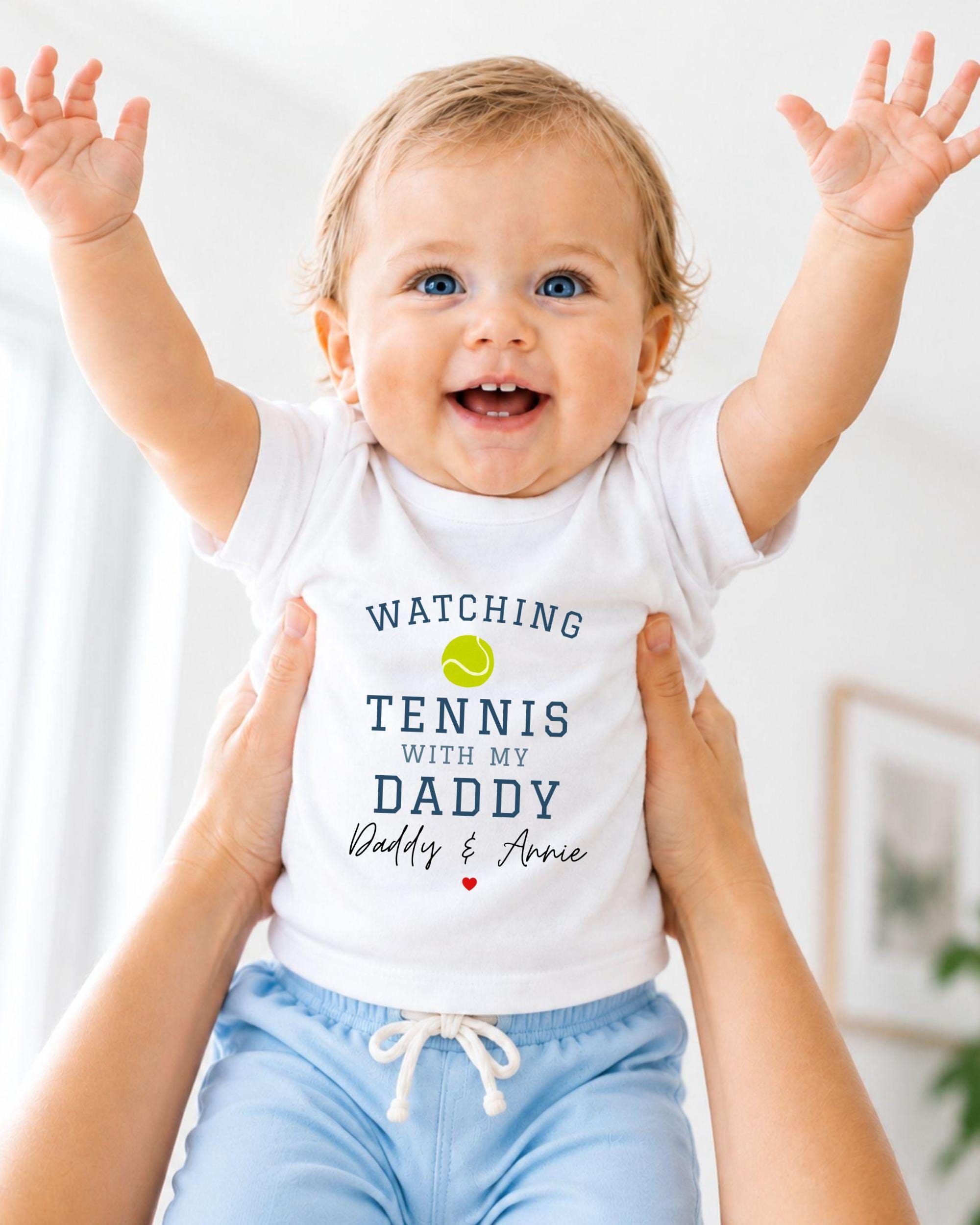 A smiling baby wearing a white shirt with the text "WATCHING TENNIS WITH MY DADDY Daddy & Annie" is being held up by someone's arms.
