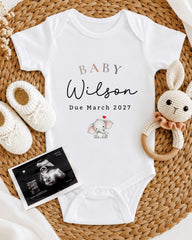 A white baby onesie with the text "BABY Wilson Due March 2027" printed on it, along with a crocheted rabbit toy and a baby bootie. There is also an Ultrasound image of a baby elephant displayed next to the onesie.