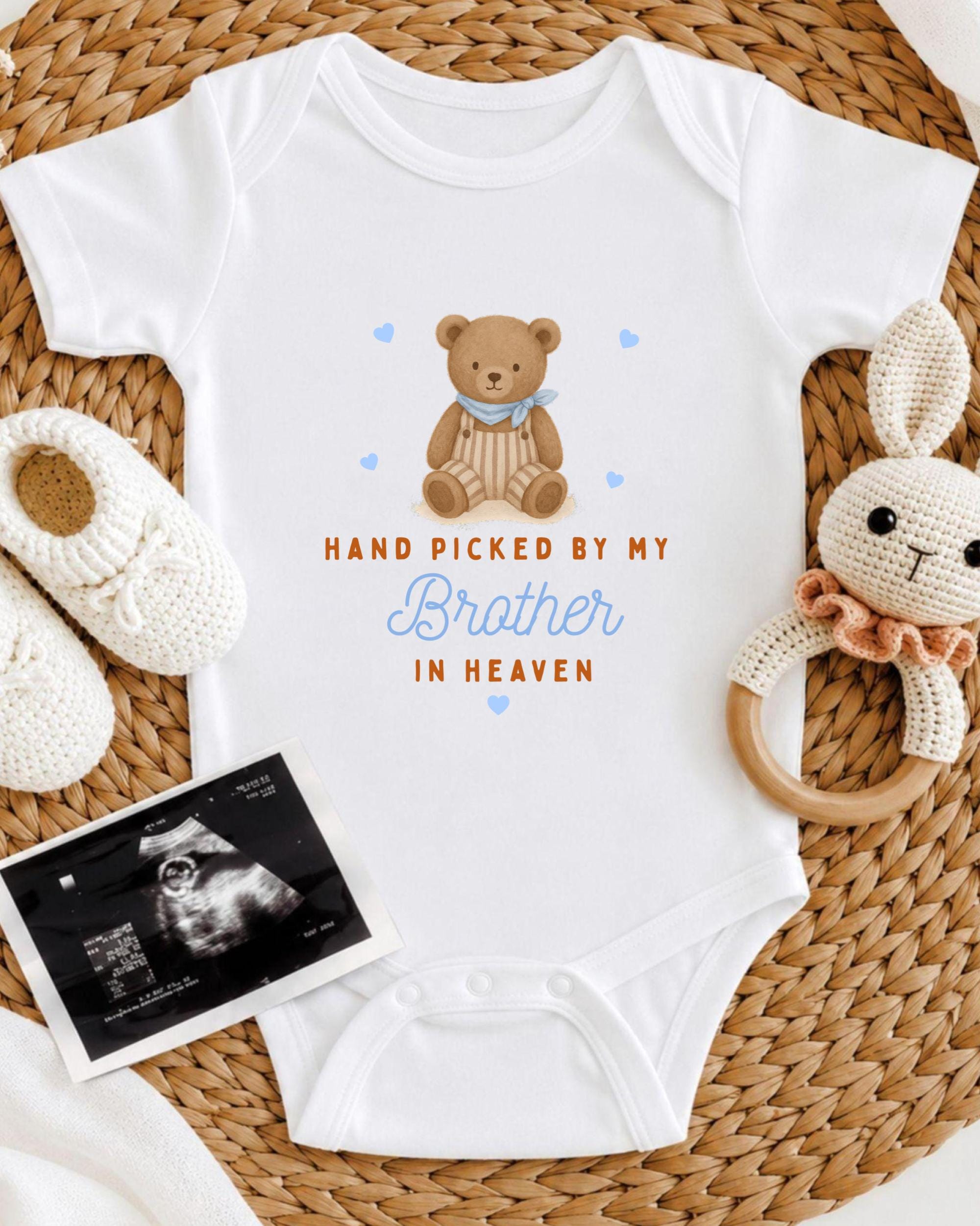 A baby onesie with a teddy bear and the text "Hand picked by my Brother in Heaven" is displayed on a woven placemat, accompanied by a crocheted bunny toy and a black and white baby photograph.