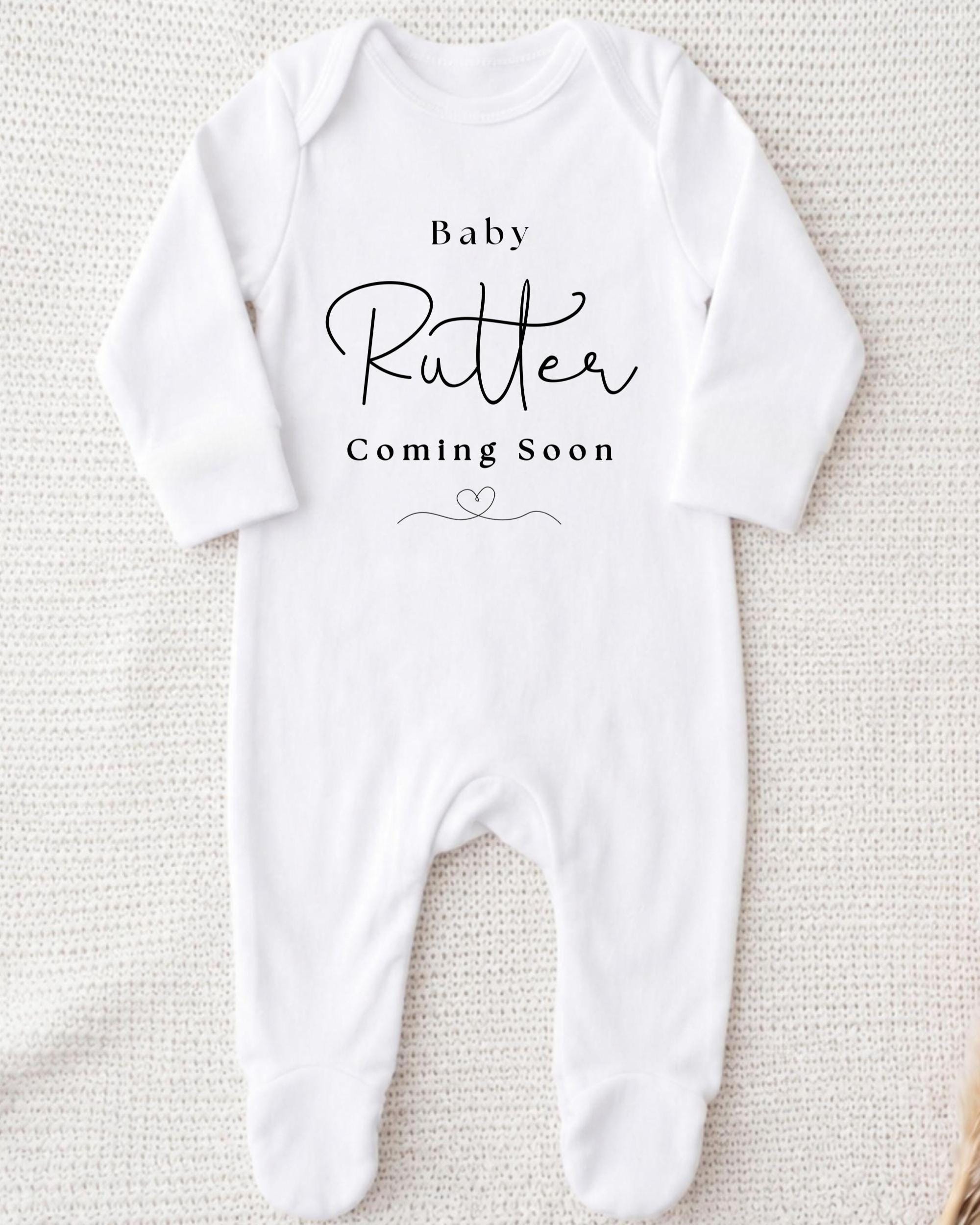 A white onesie with the text "Baby Rutter Coming Soon" printed on it.