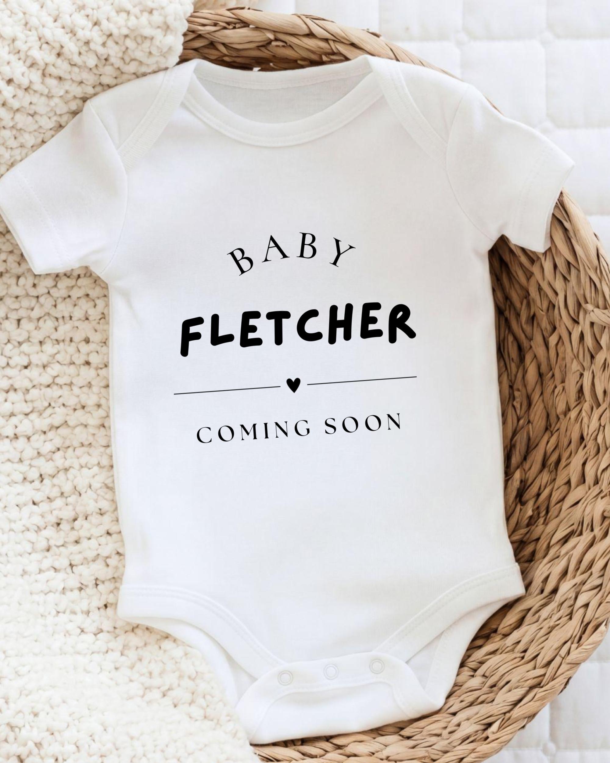 A white baby onesie with the text "BABY FLETCHER COMING SOON" printed on it, placed on a woven basket.
