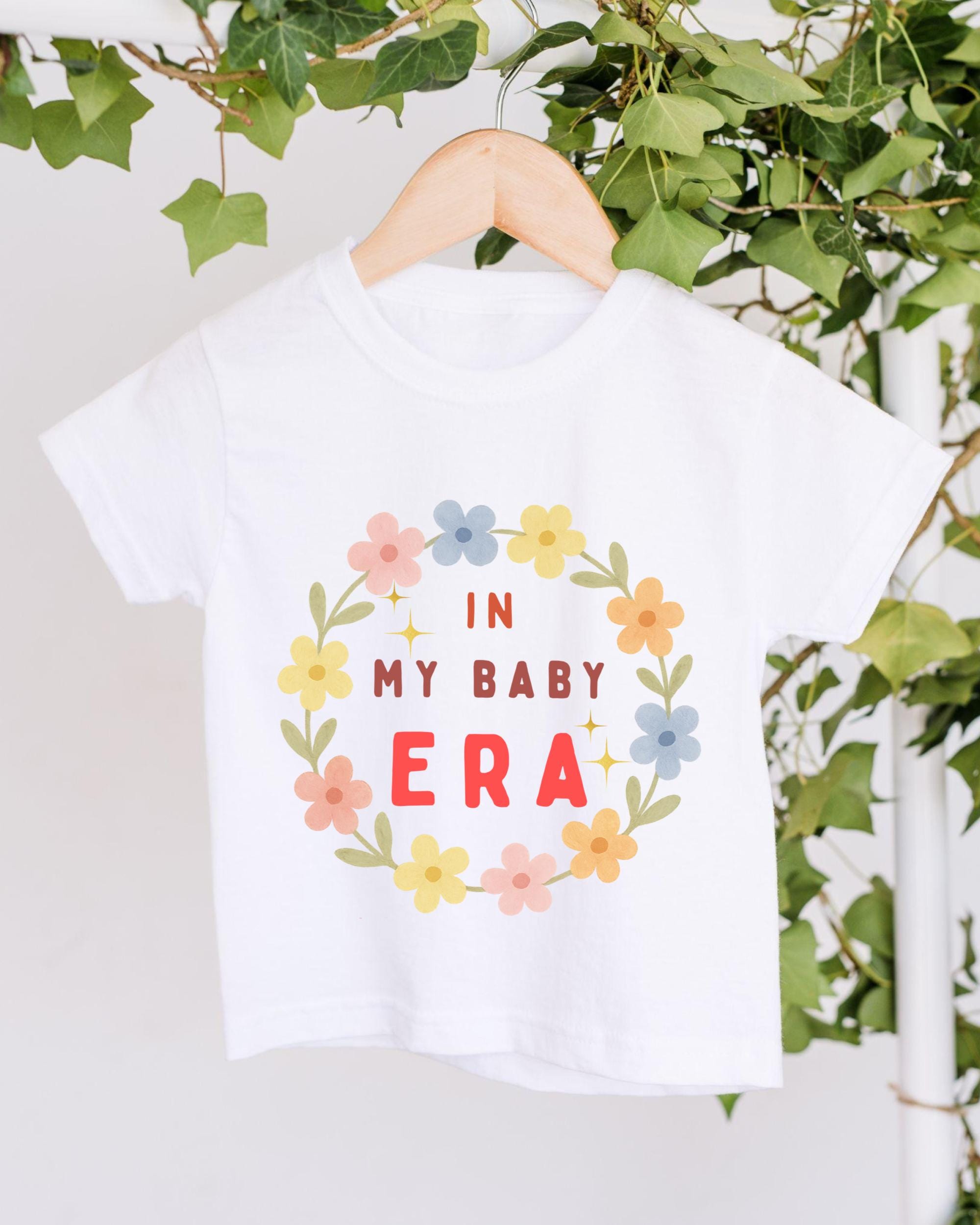 A white t-shirt with a floral wreath design and text printed on it, hanging on a wooden hanger.
