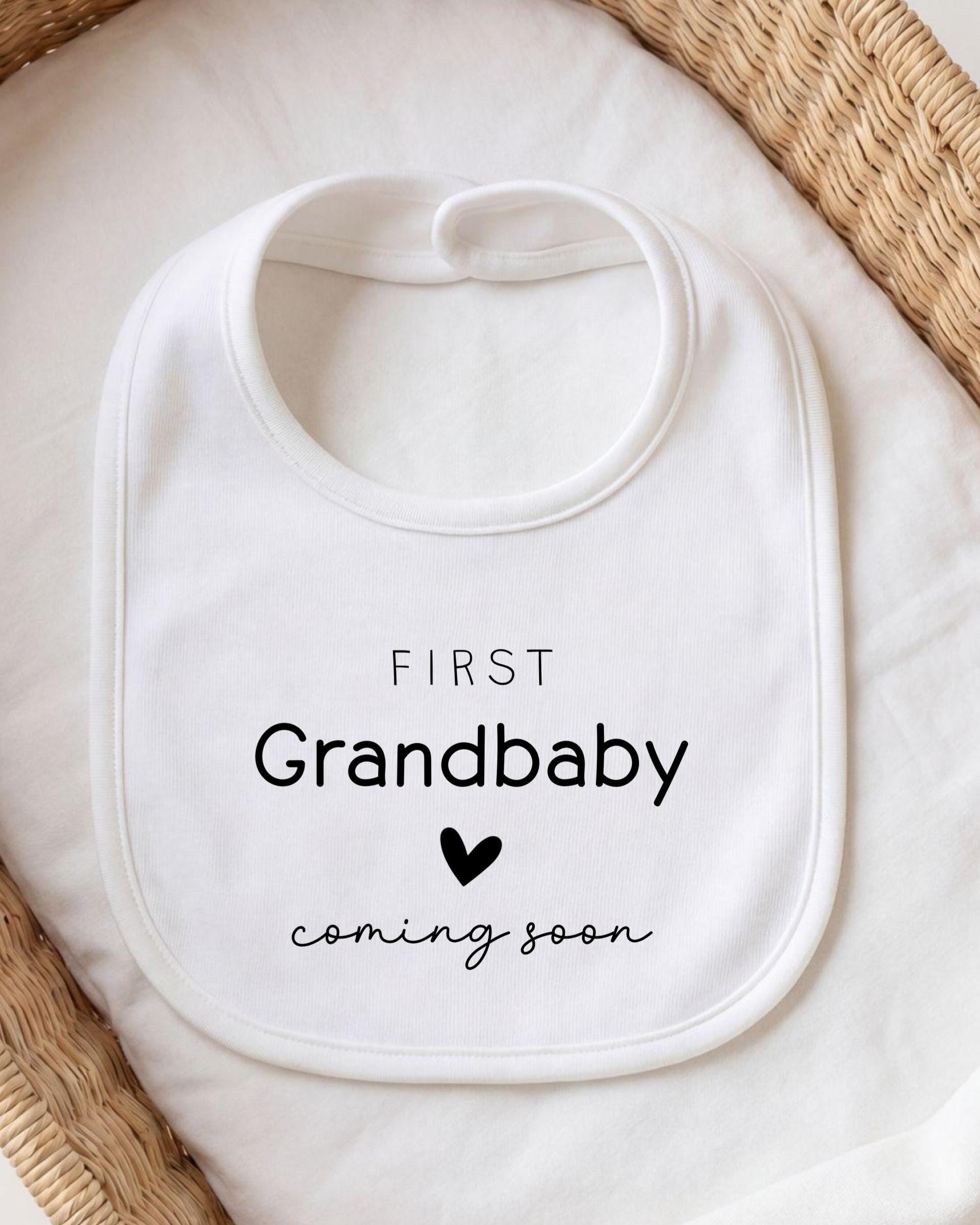 A white bib with the text "FIRST Grandbaby coming soon" printed on it, placed on a white cloth.