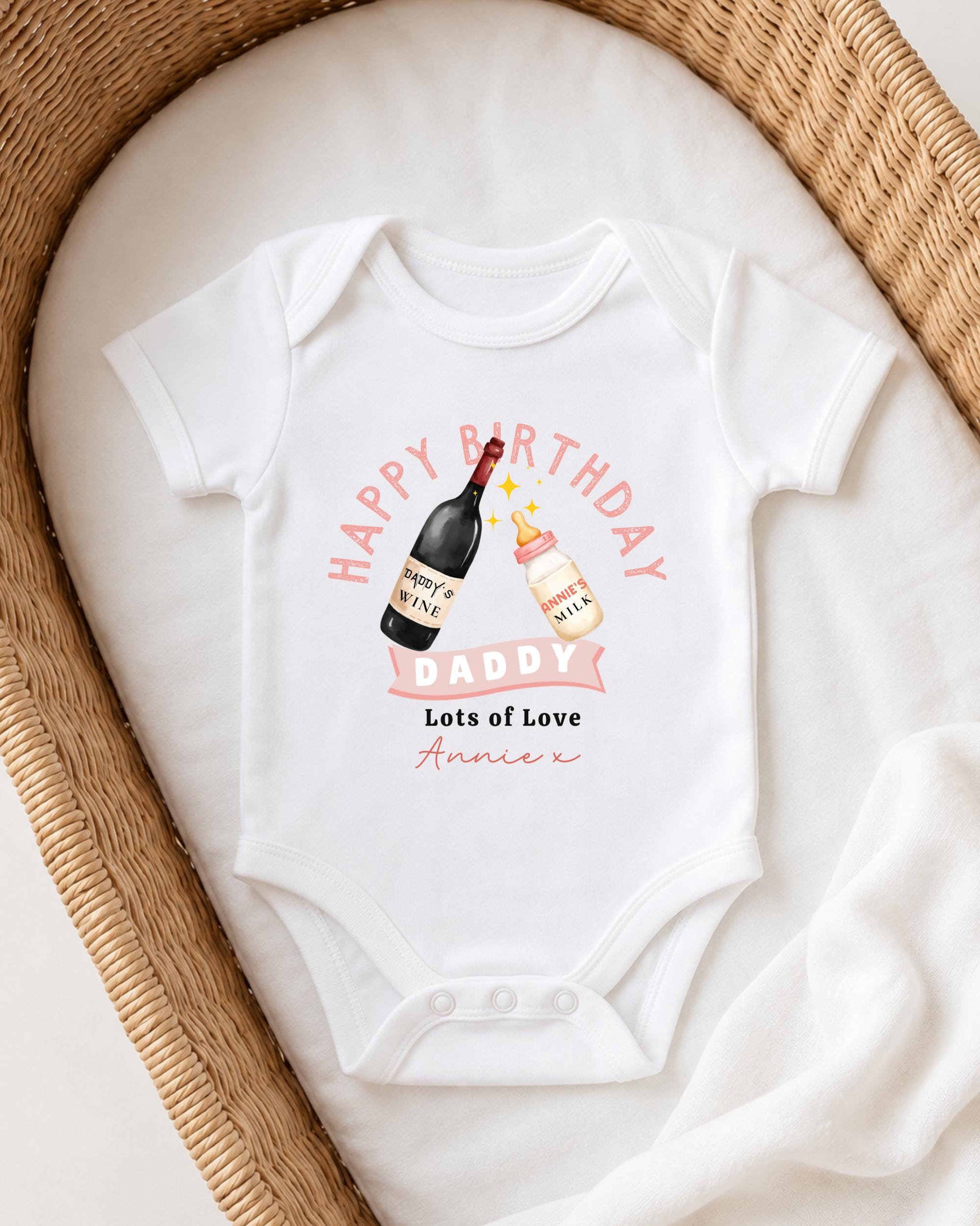 A white onesie with a birthday message and illustrations of a wine bottle and a baby bottle, placed on a white blanket in a woven basket.