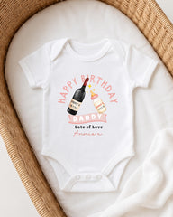 A white onesie with a birthday message and illustrations of a wine bottle and a baby bottle, placed on a white blanket in a woven basket.