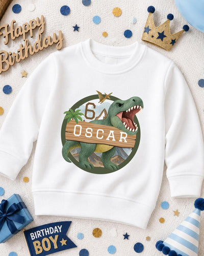 A white sweatshirt with a dinosaur graphic and the number 6, along with a birthday hat and other party decorations, suggesting a birthday celebration.