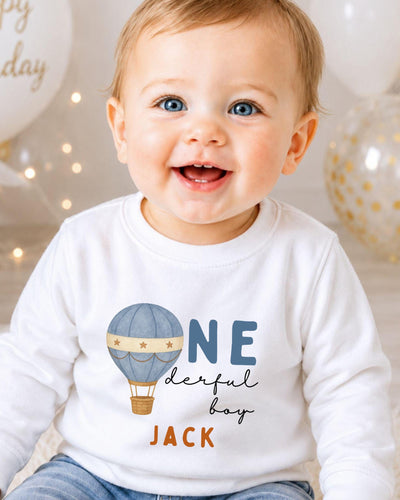 A smiling baby boy wearing a white shirt with a hot air balloon design and the text "ONE derful boy JACK" printed on it.