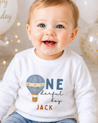 A smiling baby boy wearing a white shirt with a hot air balloon design and the text "ONE derful boy JACK" printed on it.