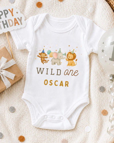 A white bodysuit with a cartoon image of a monkey, elephant, and lion, along with the text "WILD one OSCAR" printed on it, is displayed on a bed with confetti.