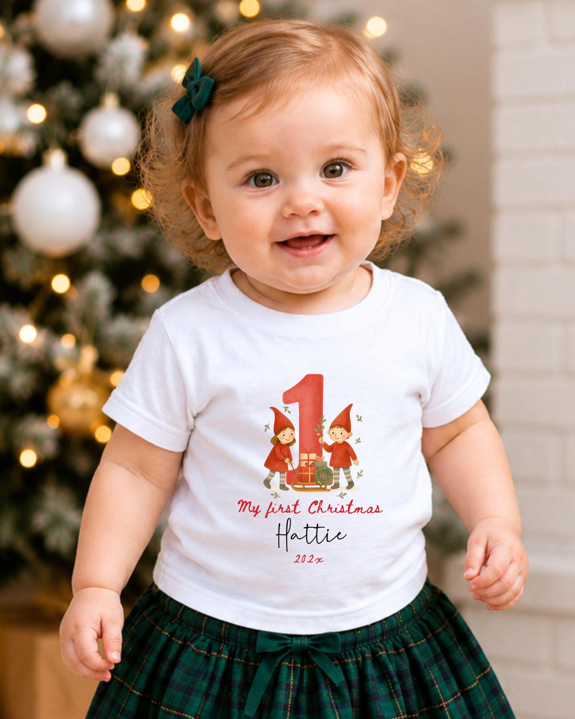 A smiling baby girl wearing a white t-shirt with a personalized number 1 and Christmas-themed design, standing in front of a Christmas tree.