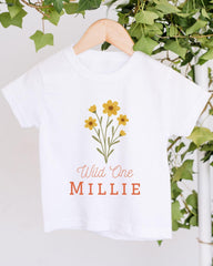 A white t-shirt with a graphic design of yellow flowers and the text "Wild One Millie" printed on it, hanging on a wooden hanger.