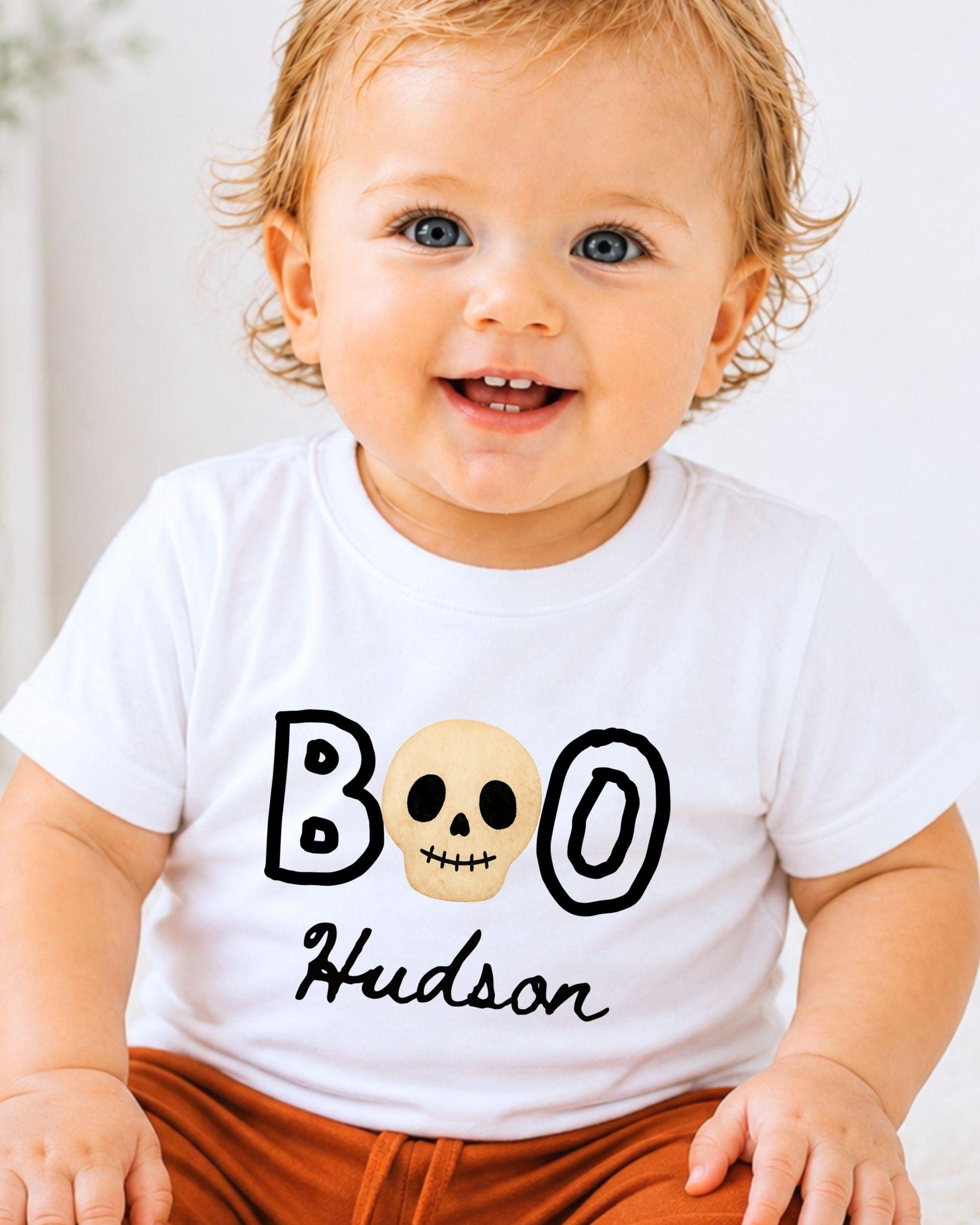 A smiling baby wearing a white t-shirt with a skull design and the text "BOO Hudson" printed on it.