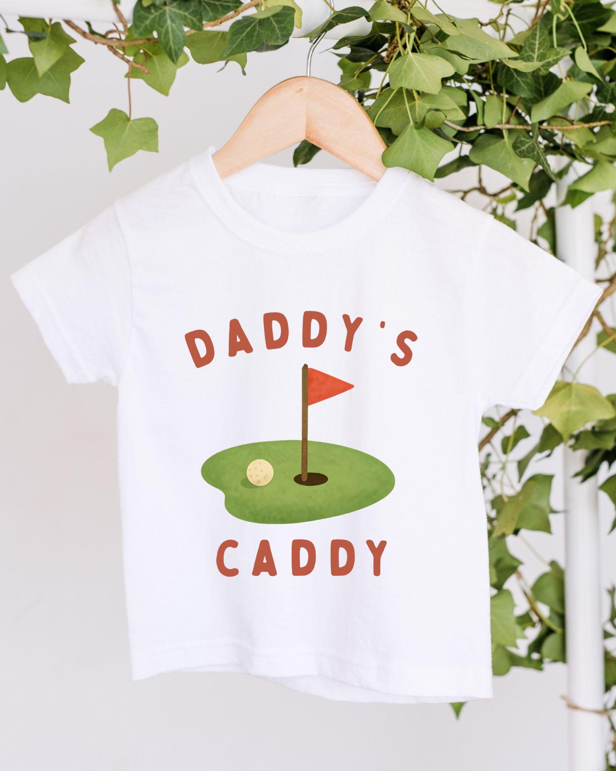 A white t-shirt with a graphic design featuring a golf hole and a flag, along with the text "DADRY'S CADDY".