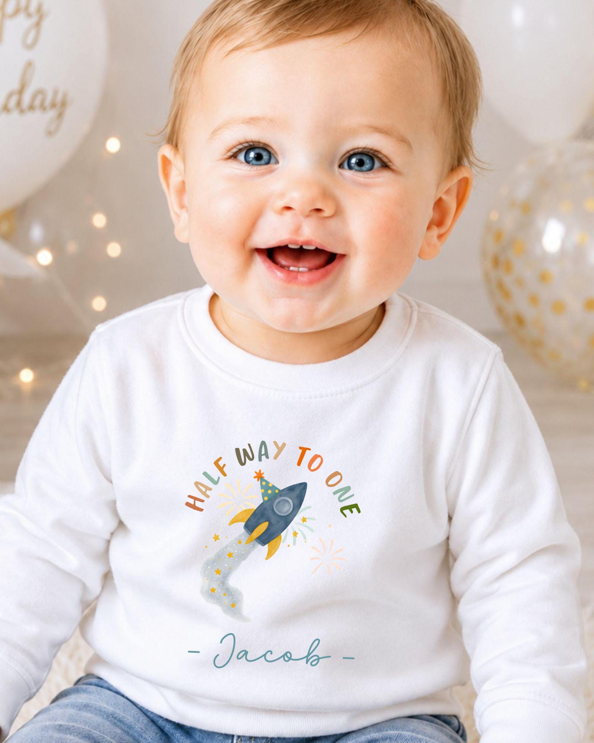 A smiling baby boy wearing a white shirt with a rocket ship graphic and the text "Half Way to One" is sitting in front of birthday balloons.