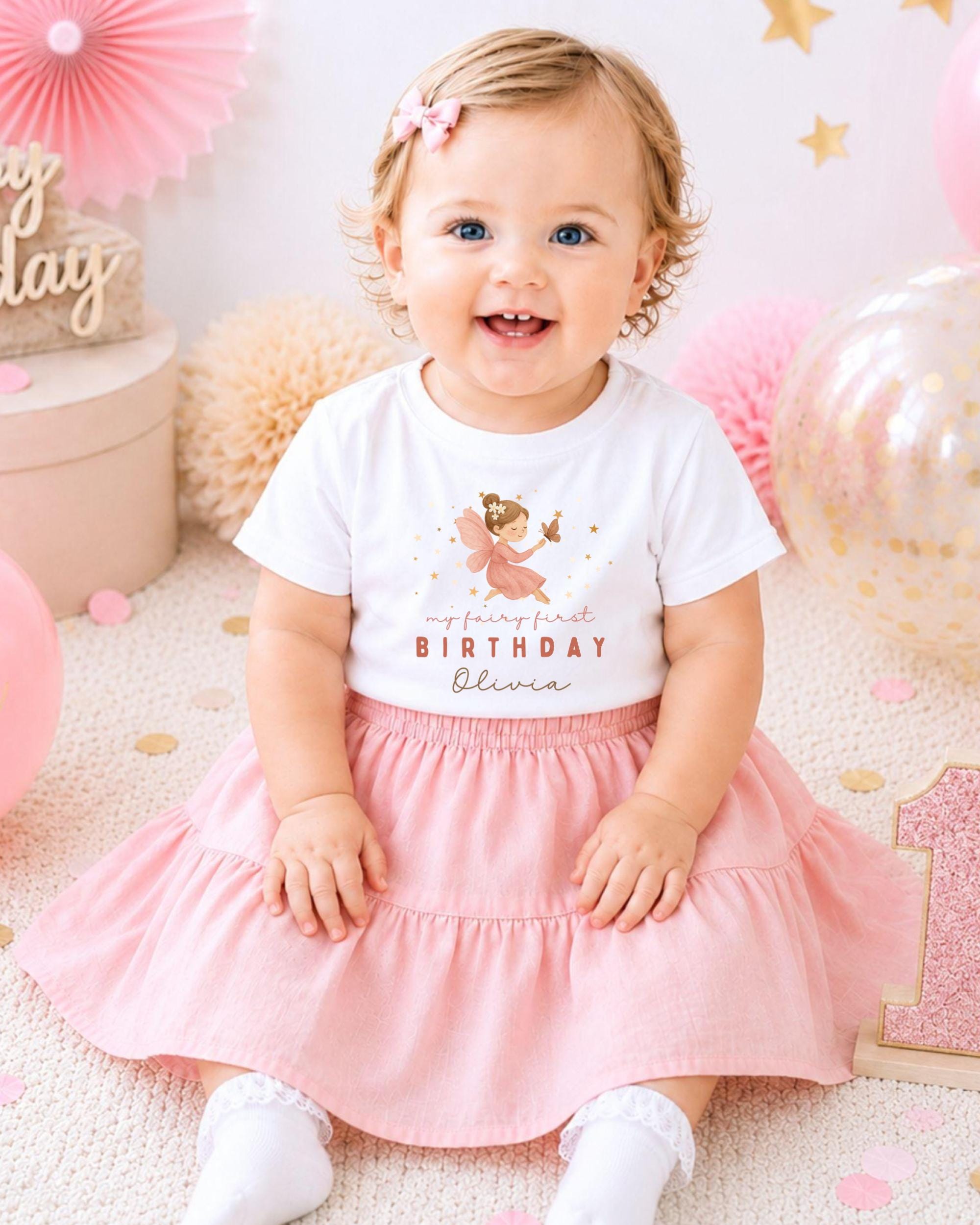 A smiling baby girl wearing a white t-shirt with a fairy design and a pink skirt, sitting on a pink and white background with balloons and confetti.