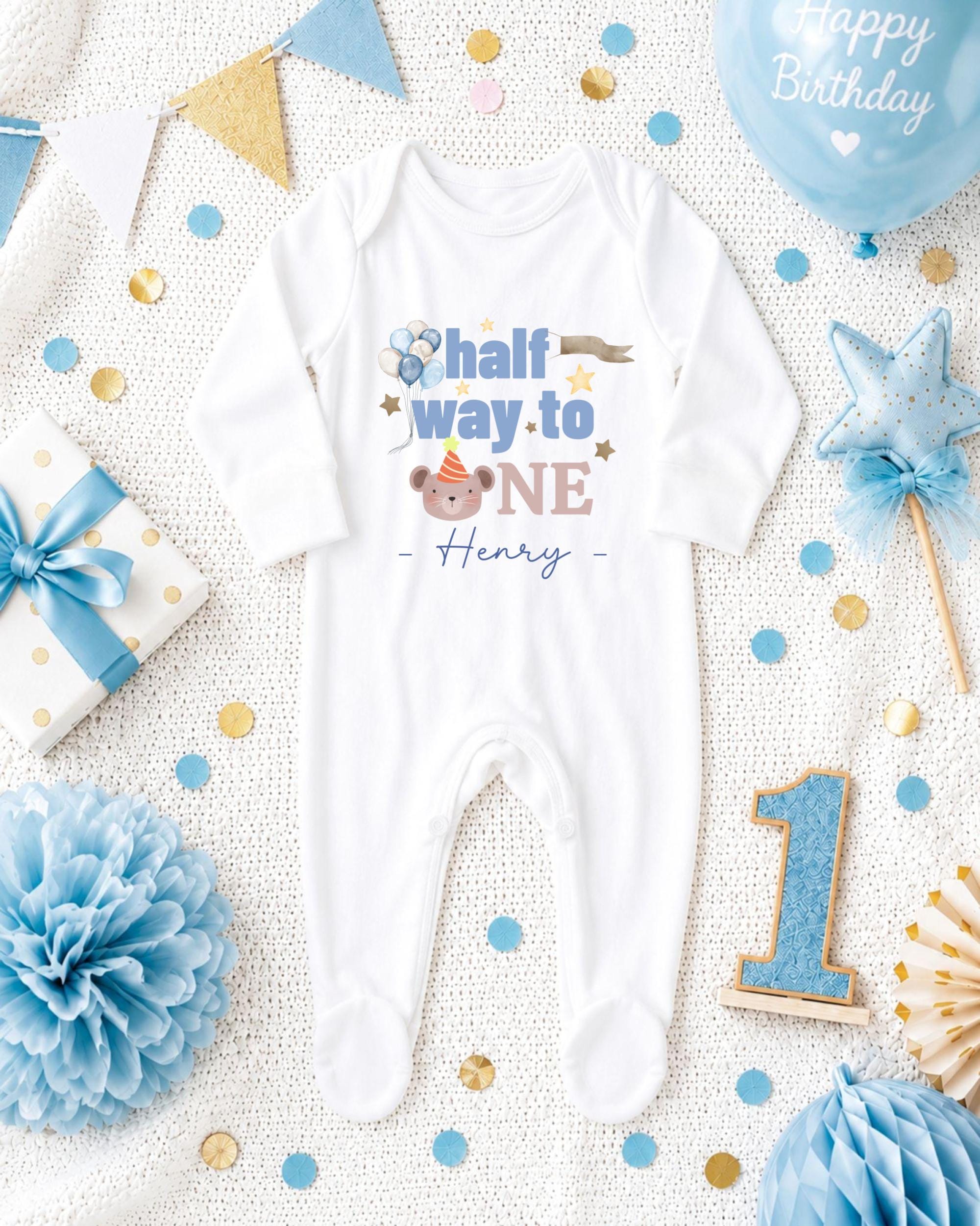 A white onesie with the text "half way to one" printed on it, surrounded by birthday decorations such as a blue balloon, blue and gold confetti, and a blue and gold star.