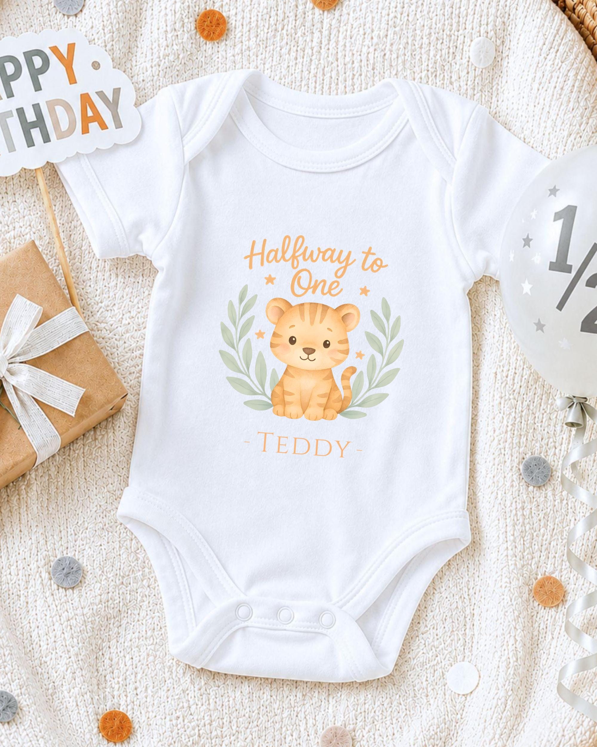 A white onesie with a cartoon tiger graphic and the text "Halfway to One Teddy" printed on it, along with a "Happy Birthday" sign and confetti.