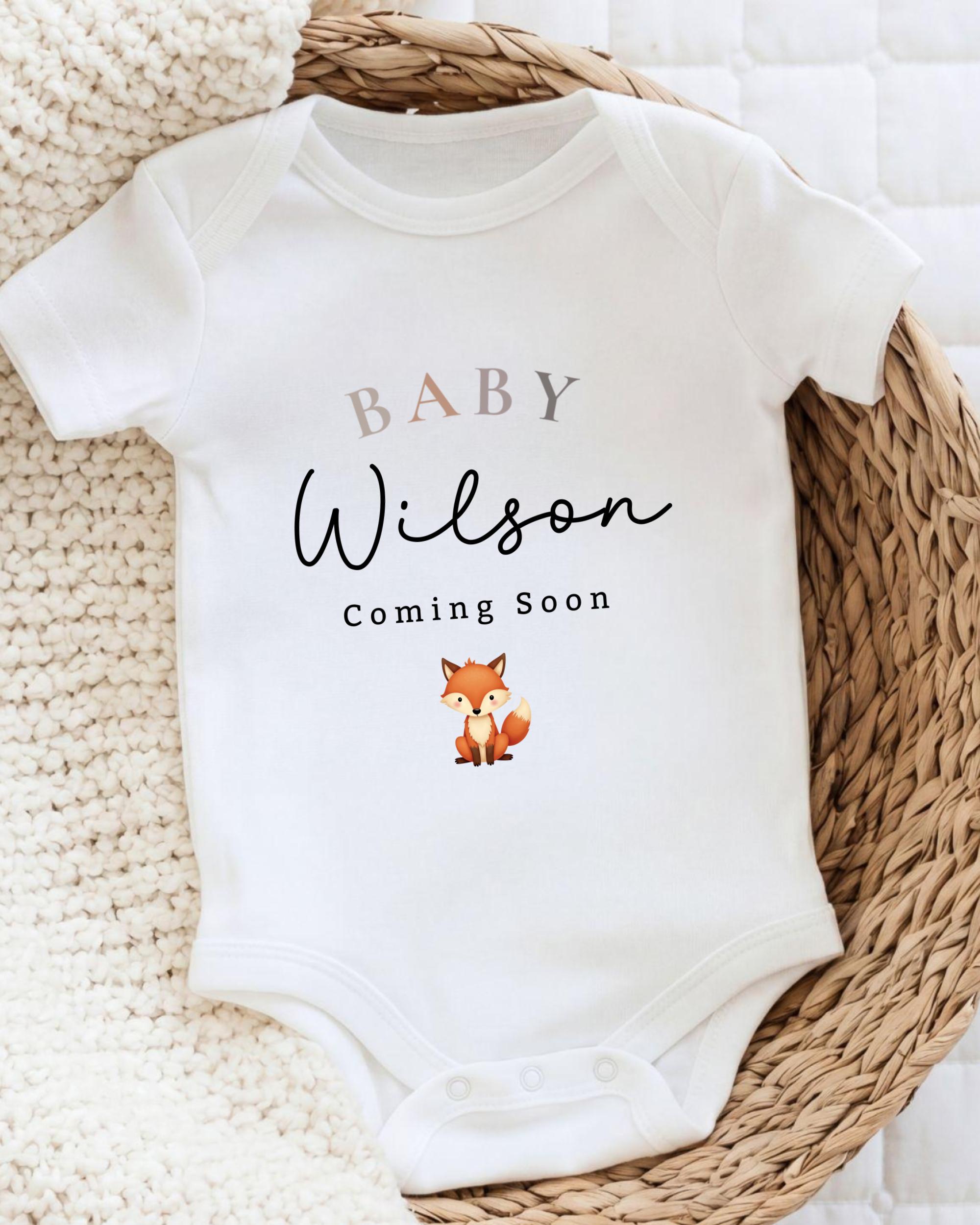 A white baby onesie with the text "BABY WILSON Coming Soon" printed on it, along with a small illustration of a fox.