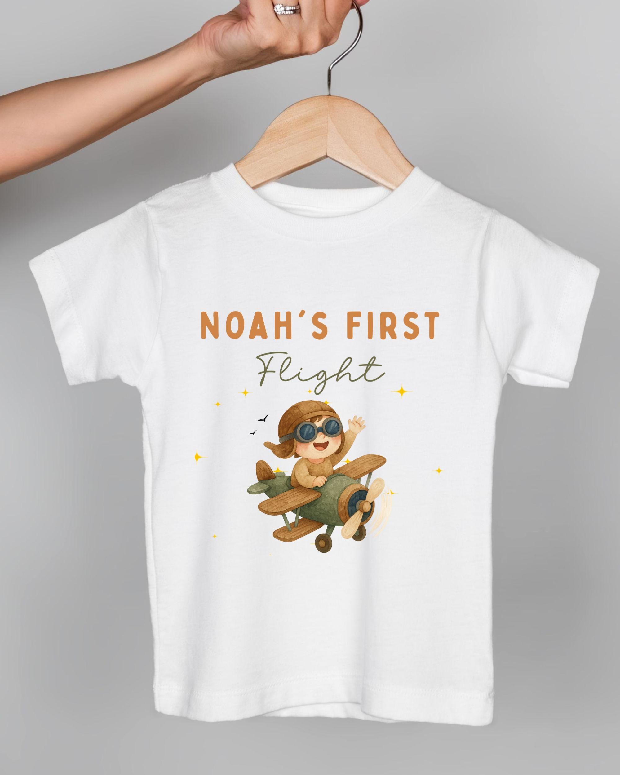 A white t-shirt with a cartoon image of a child wearing a pilot's hat and flying a small airplane, along with the text "Noah's first flight" printed on it.