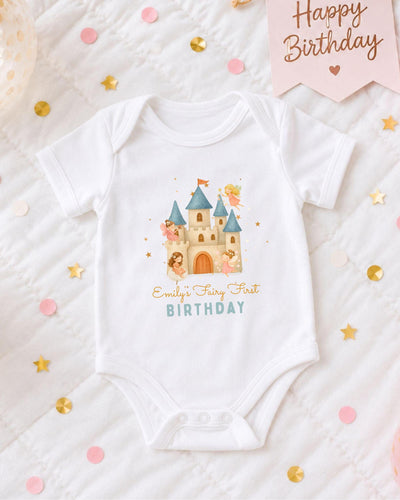 A white bodysuit with a castle design and the text "Emilly's Fairy First Birthday" printed on it, along with confetti and a birthday banner.